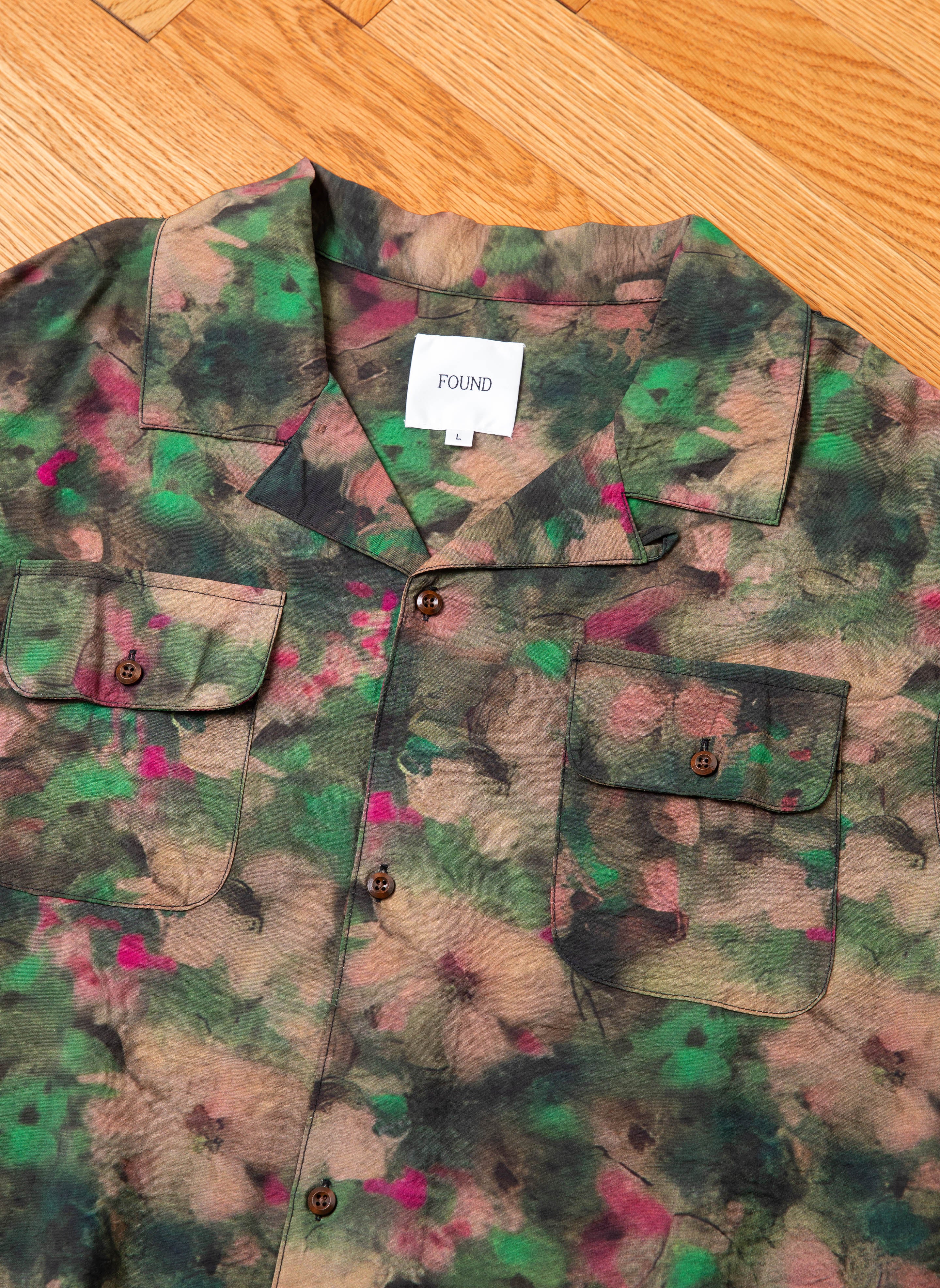 A Charlot LS Camp Shirt in camo print featuring green, brown, and pink hues lies flat on a wooden floor. This stylish shirt from the brand FOUND includes dual chest pockets and button closures, seamlessly blending functionality with fashion. A label inside the collar reads "FOUND.