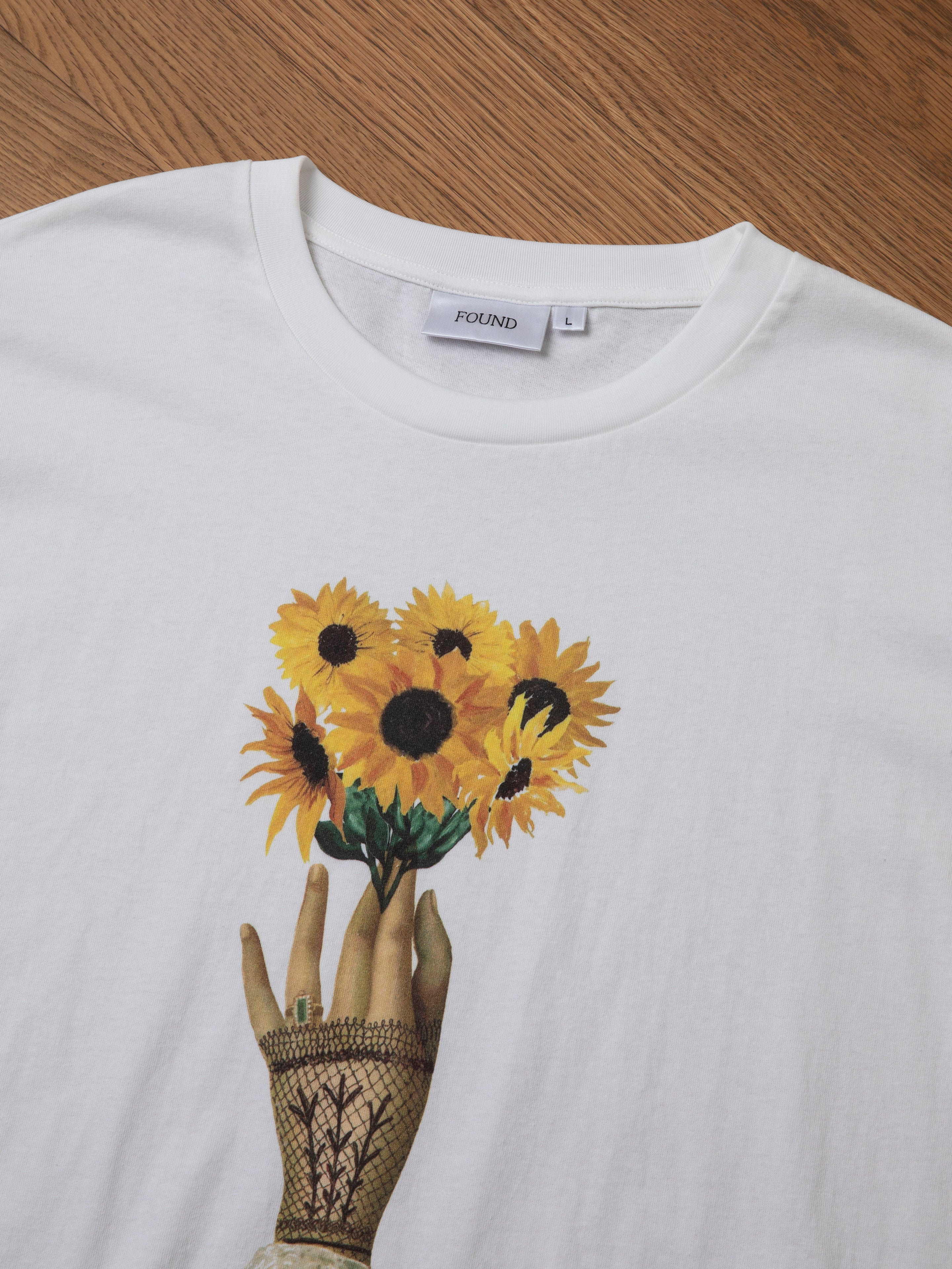 The FOUND Sunflower Tee showcases a floral graphic of a hand holding a bouquet of sunflowers against a wooden surface, offering a charming vintage feel.