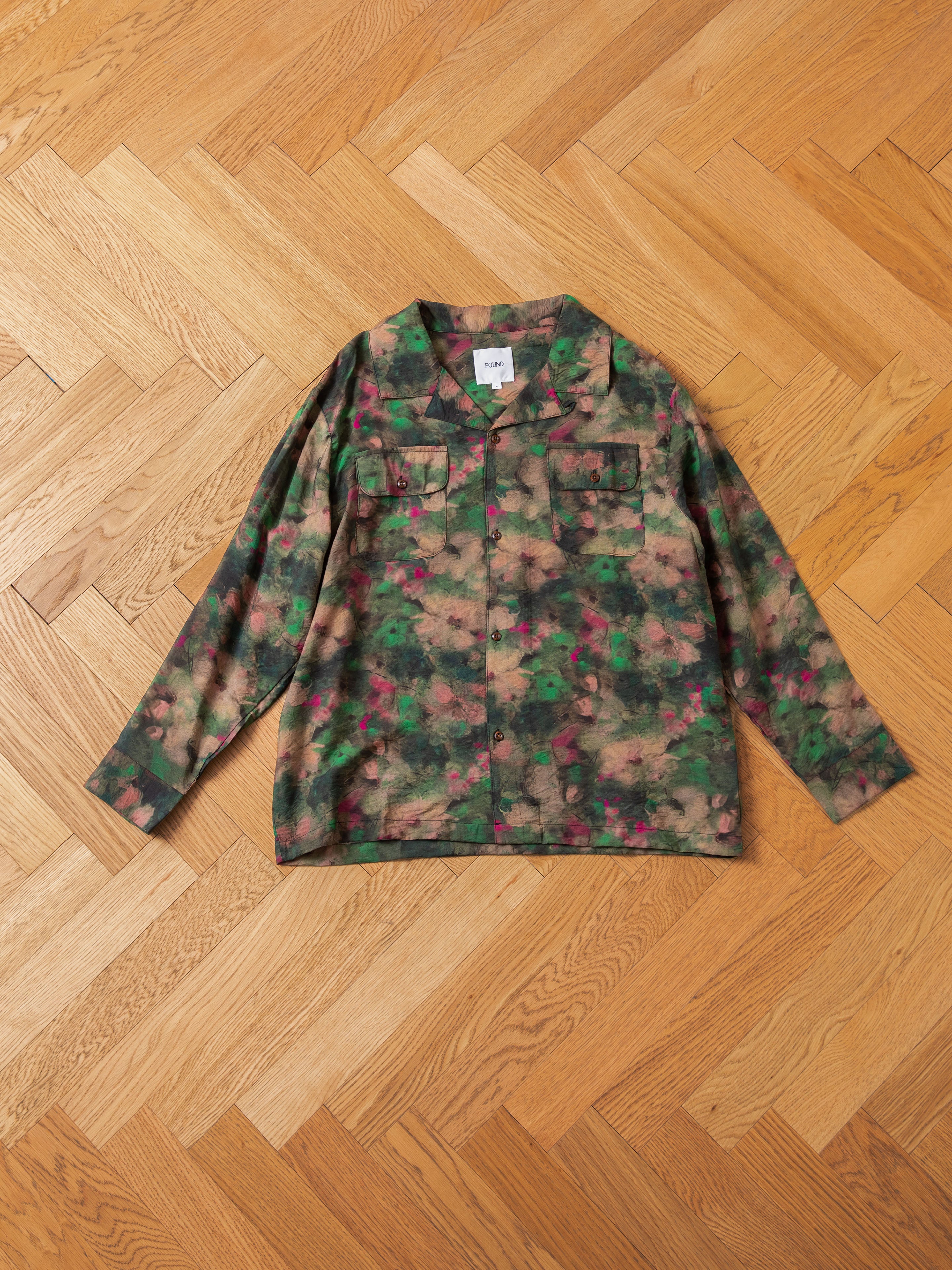 The FOUND Charlot LS Camp Shirt, showcasing a camouflage pattern and dual chest pockets, is laid flat on a wooden herringbone floor.