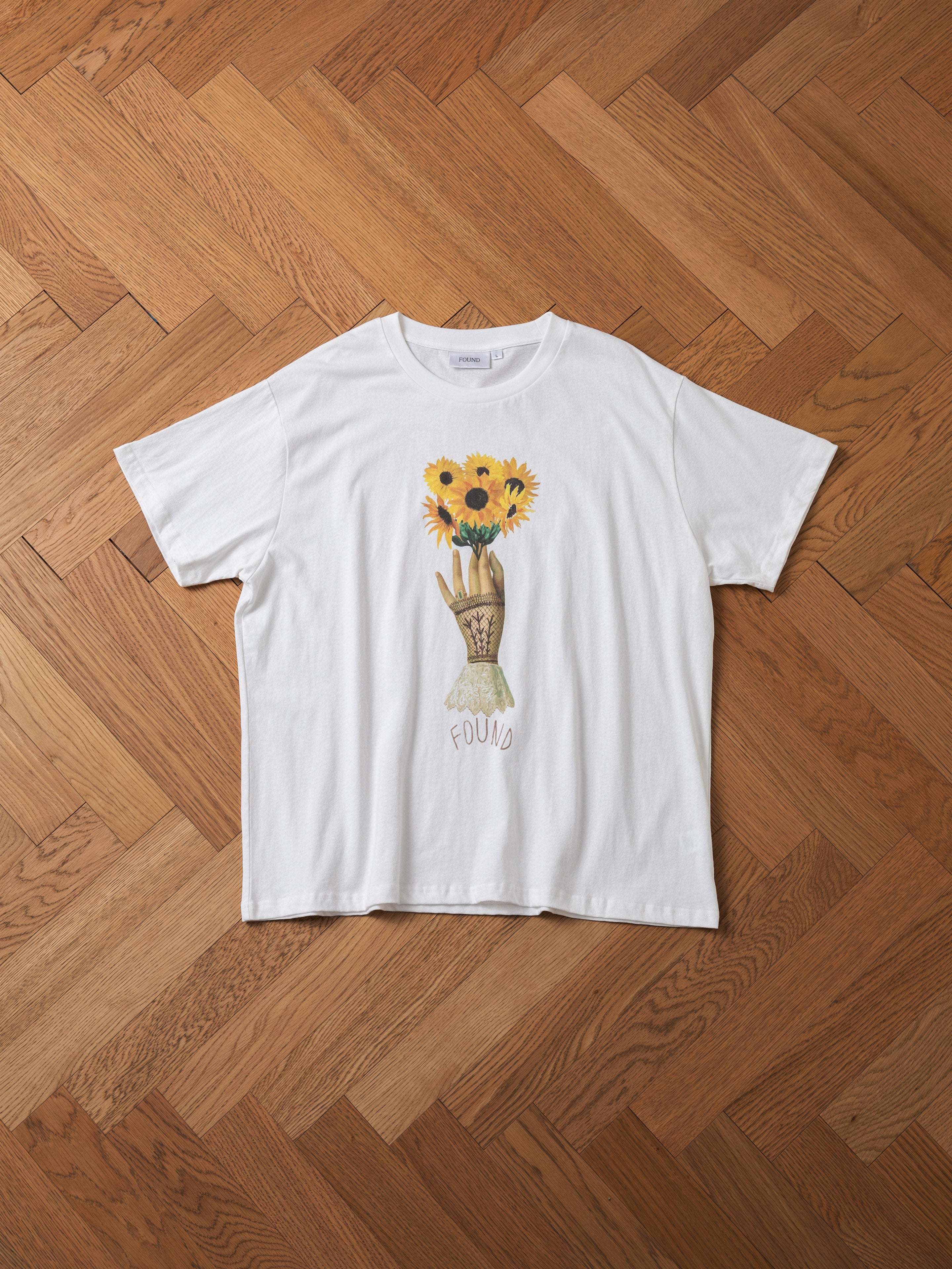 A Sunflower Tee from the brand FOUND, adorned with a floral graphic depicting sunflowers in a vase and the word "FOUND" beneath the illustration, laid flat on a wooden herringbone-patterned floor for a vintage aesthetic.