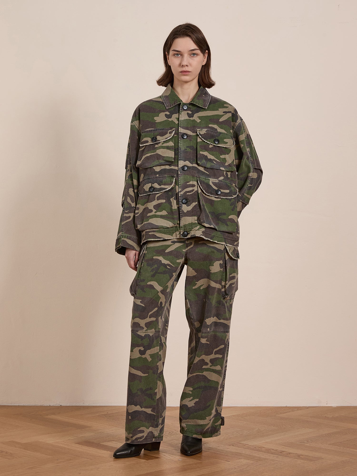 Against a plain beige background, a person stands on a wooden floor wearing FOUND's Camouflage Twill Utility Cargo Pants and a loose-fitting camouflage jacket, exuding a vintage look.