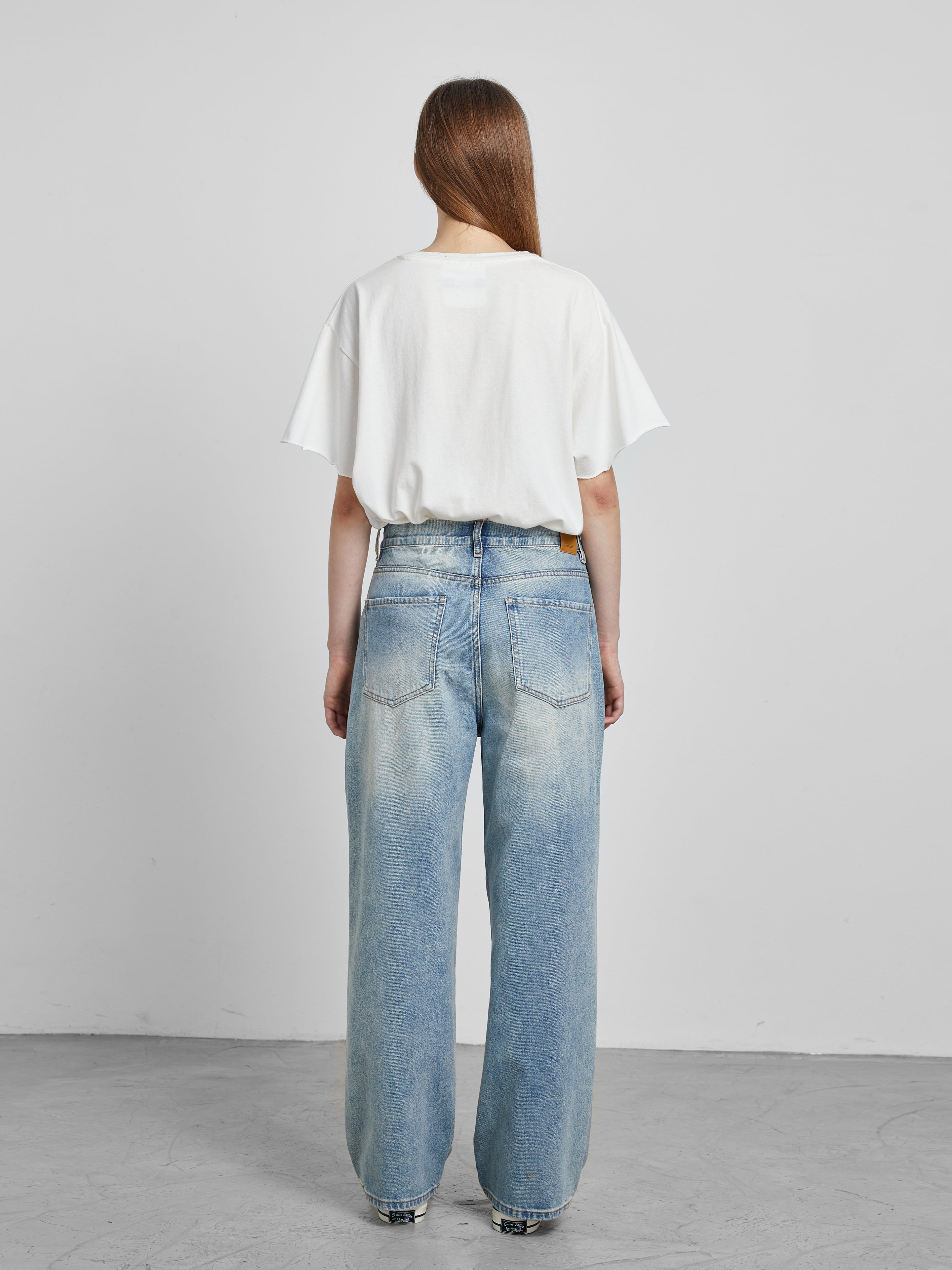 A person with long hair faces away, wearing a white t-shirt and Mix Patch Jeans by Profound in a minimalist setting.