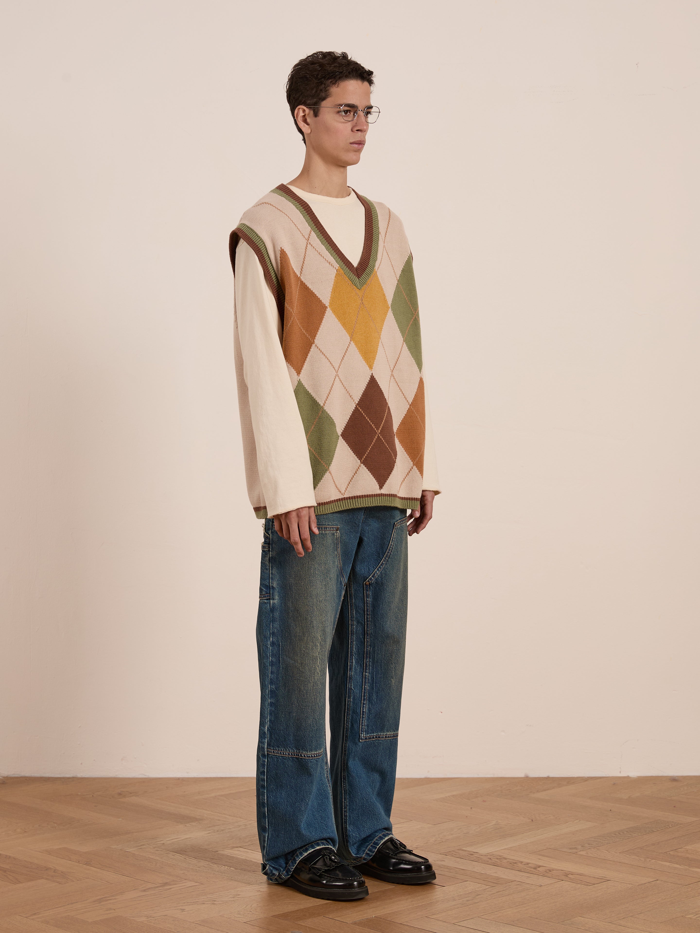 A person showcases the FOUND Argyle Knit Vest in beige with a vintage-style diamond pattern, paired with a long-sleeve shirt, blue jeans, and black shoes on a plain backdrop. The unisex relaxed fit of the vest effortlessly completes the look.