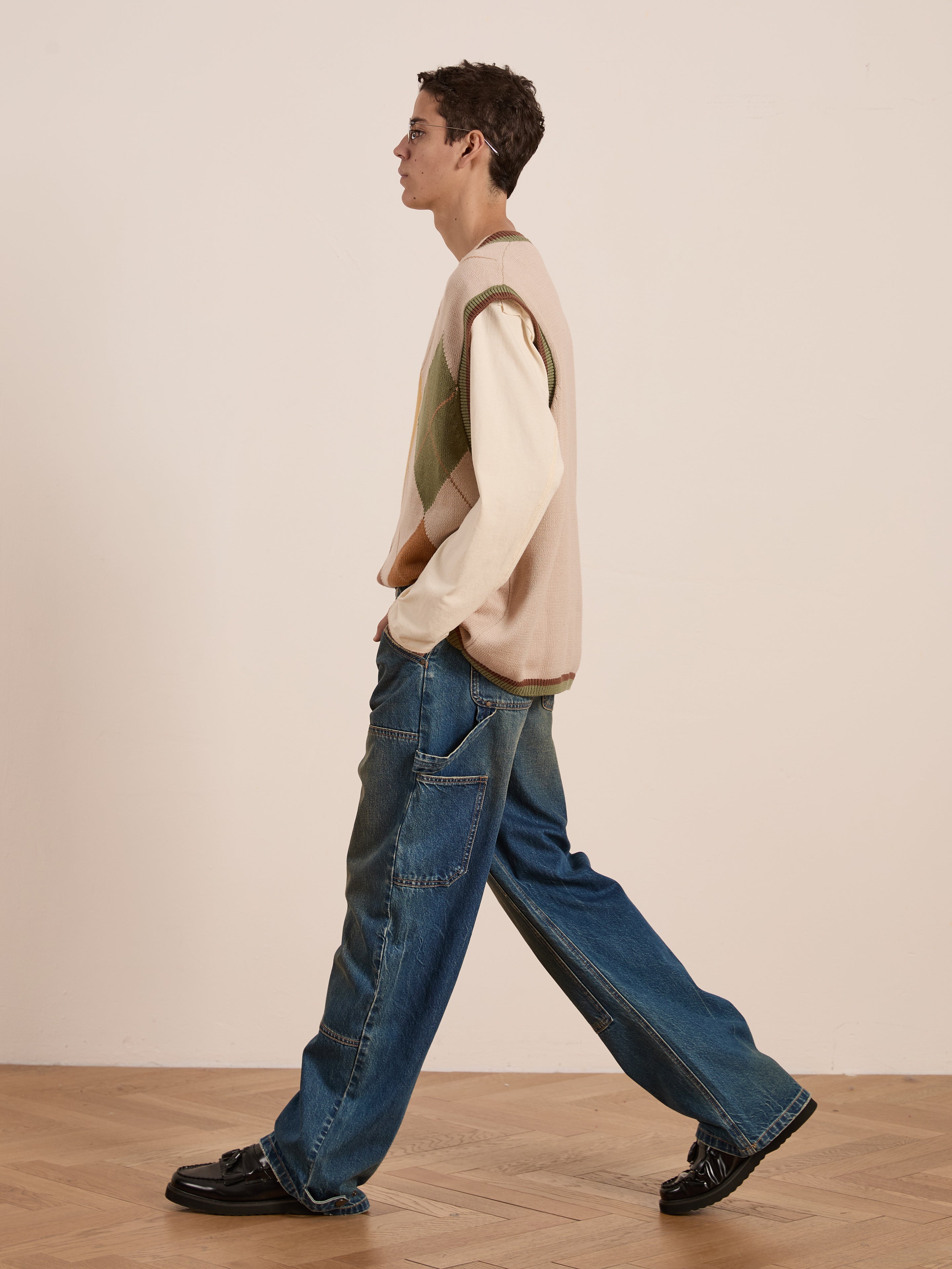 A person walks on the wooden floor wearing FOUND's Argyle Knit Vest, a vintage-style diamond pattern piece, over a long-sleeve shirt, paired with unisex relaxed fit baggy jeans and black shoes.