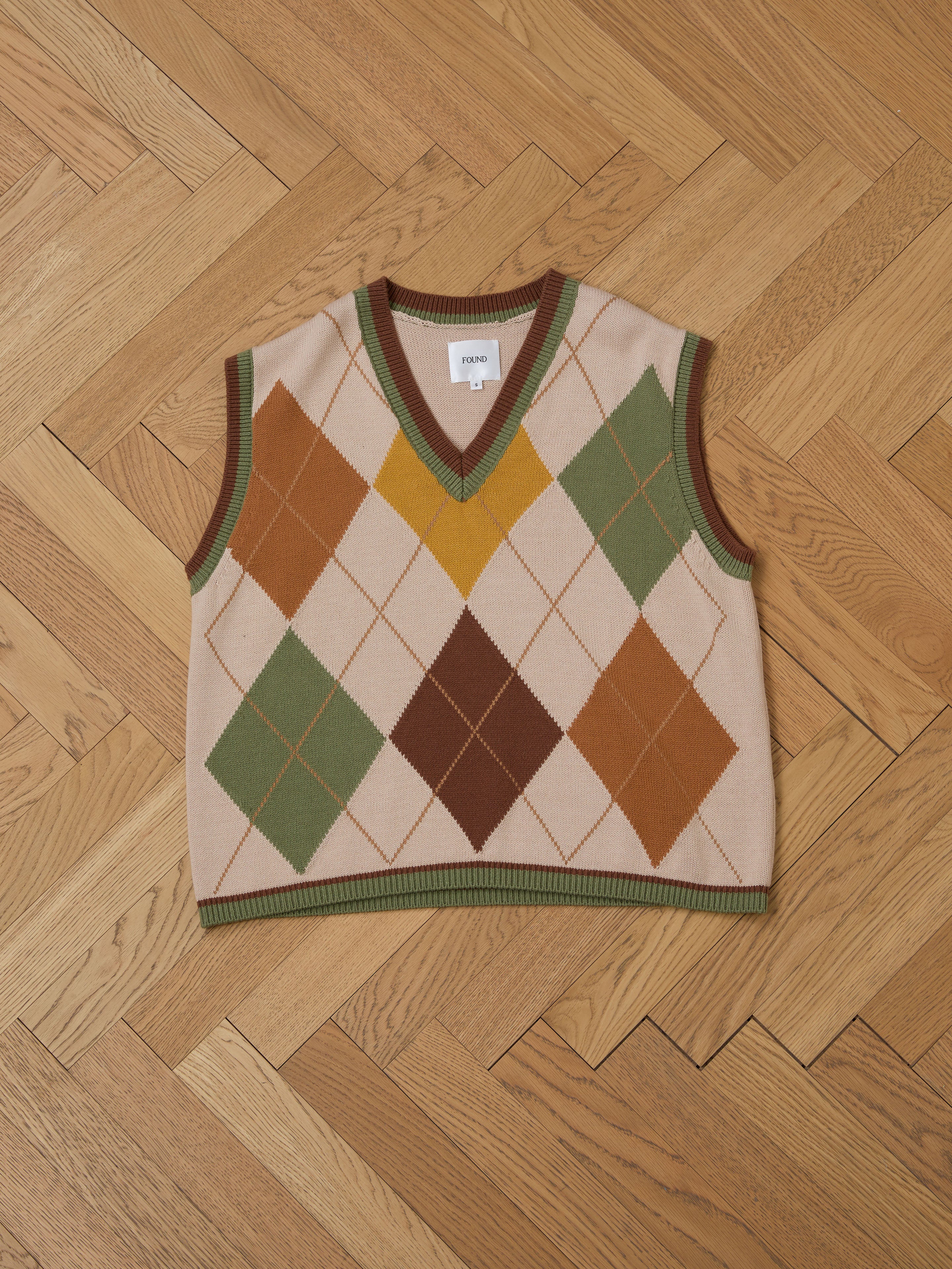 The FOUND Argyle Knit Vest features a vintage diamond pattern in green, brown, and orange hues on a wooden herringbone floor, offering a unisex relaxed fit for a timeless wardrobe addition.