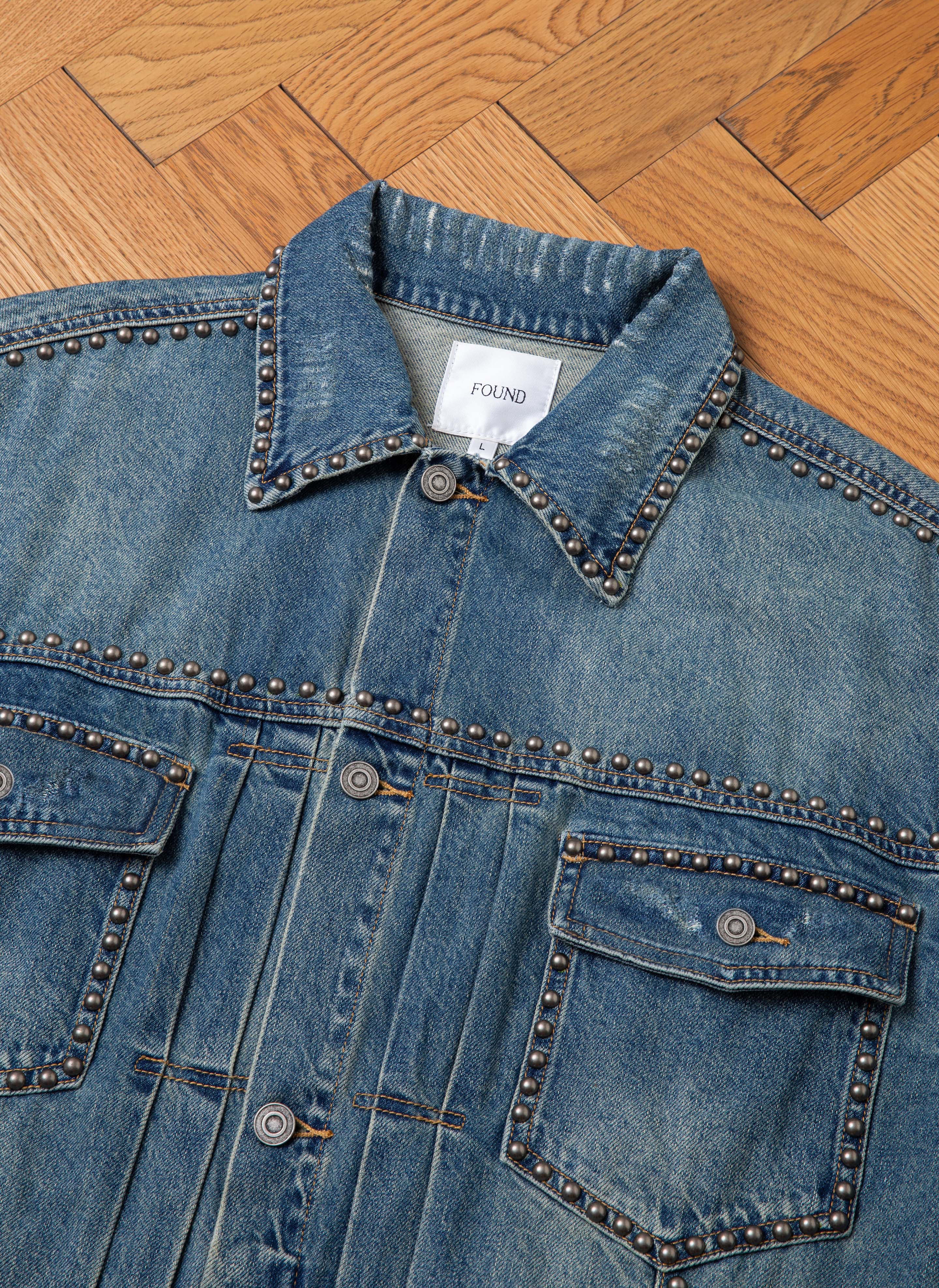 Displayed on a wooden floor, the Azar Studded Denim Trucker Jacket from FOUND showcases a vintage blue wash with an oversized boxy fit. Metal stud accents adorn the collar, placket, and pockets, while the label inside the collar proudly bears the "FOUND" brand name.