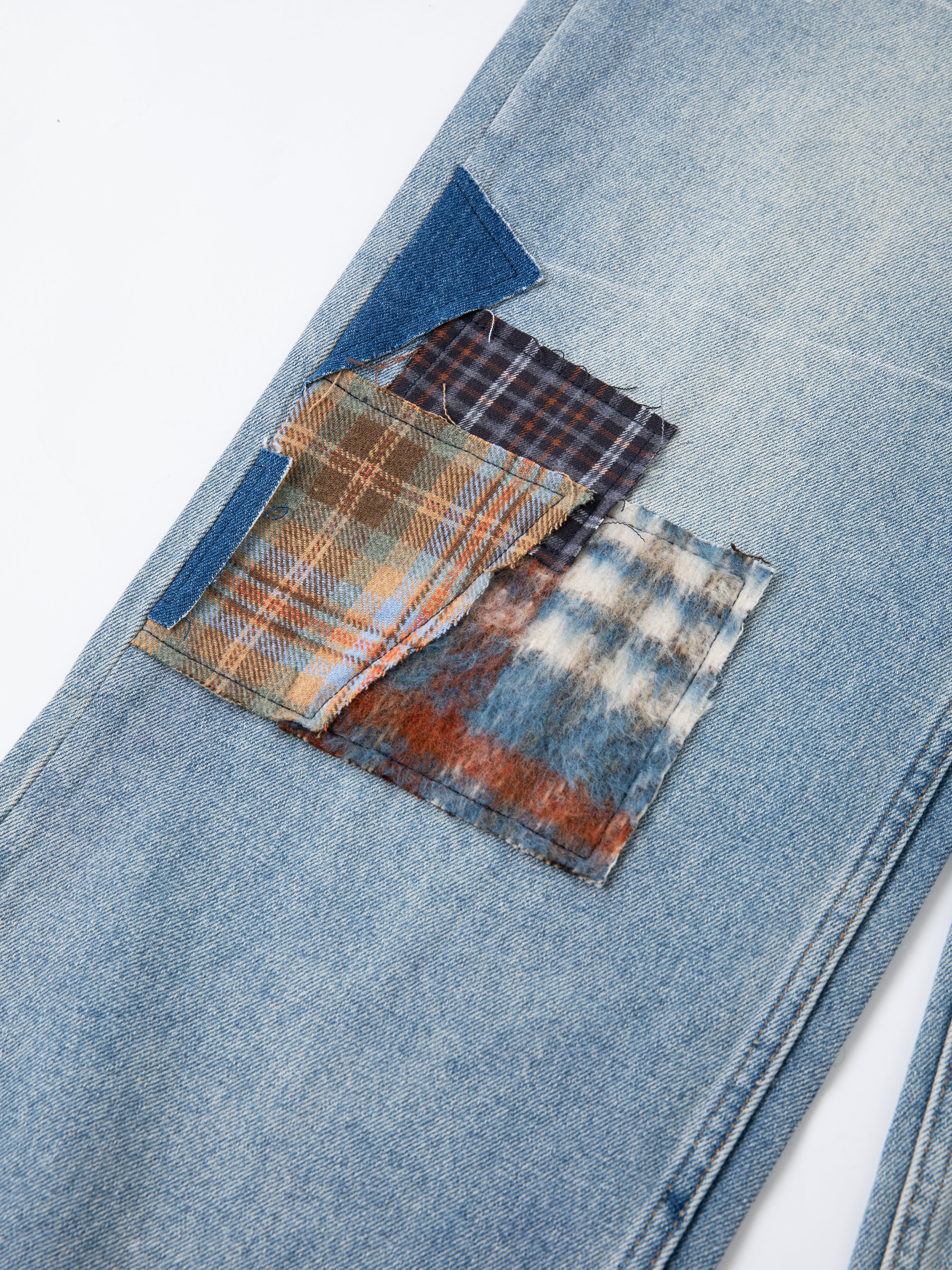 A close-up of Profound's Mix Patch Jeans displays four overlapping plaid fabric patches sewn onto distressed light blue denim cotton, creating a vintage patchwork look.