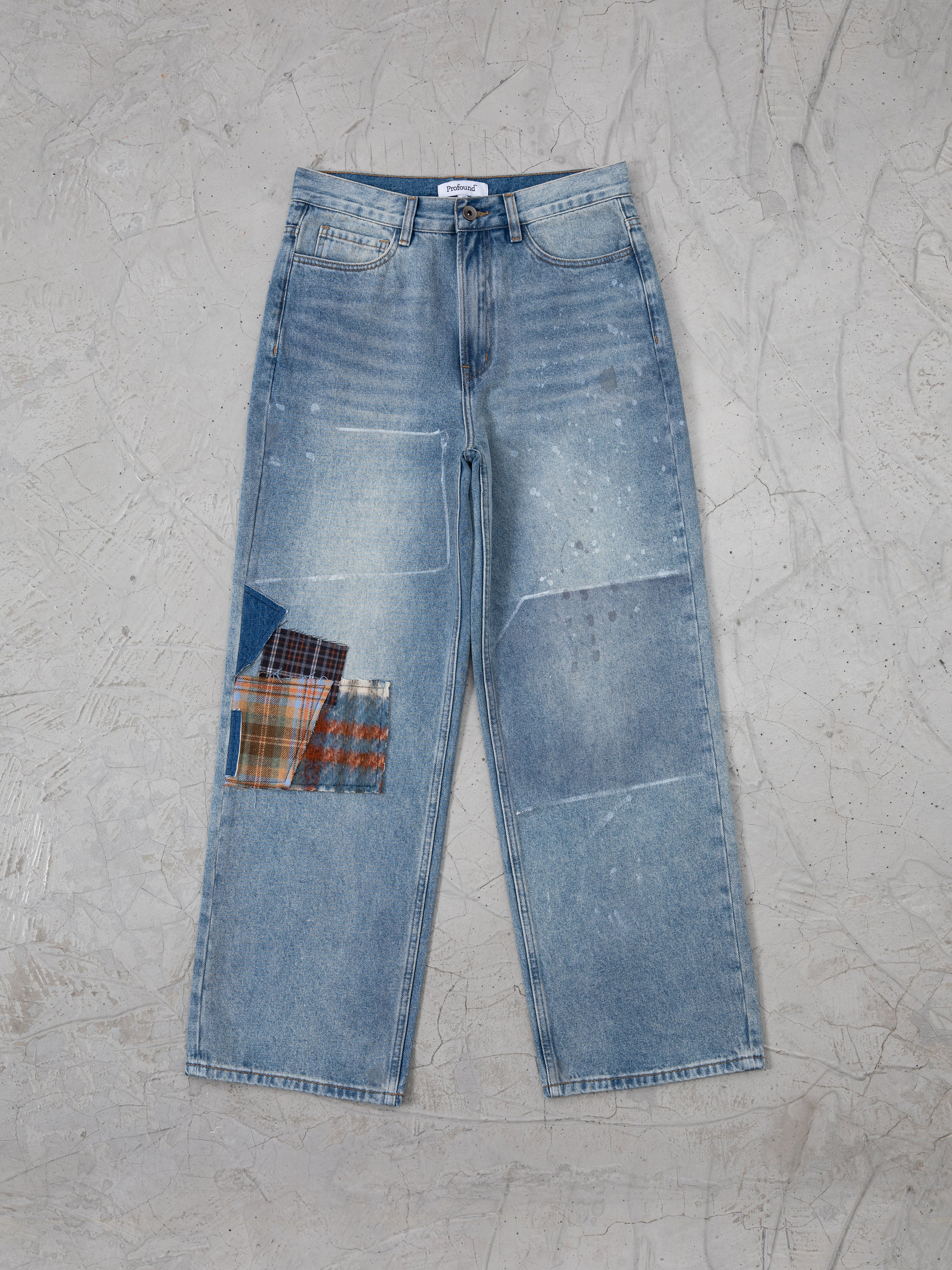 Profound's Mix Patch Jeans, featuring a faded blue vintage style with wide legs and a vibrant patchwork design on the right thigh, are laid flat on a textured gray surface.