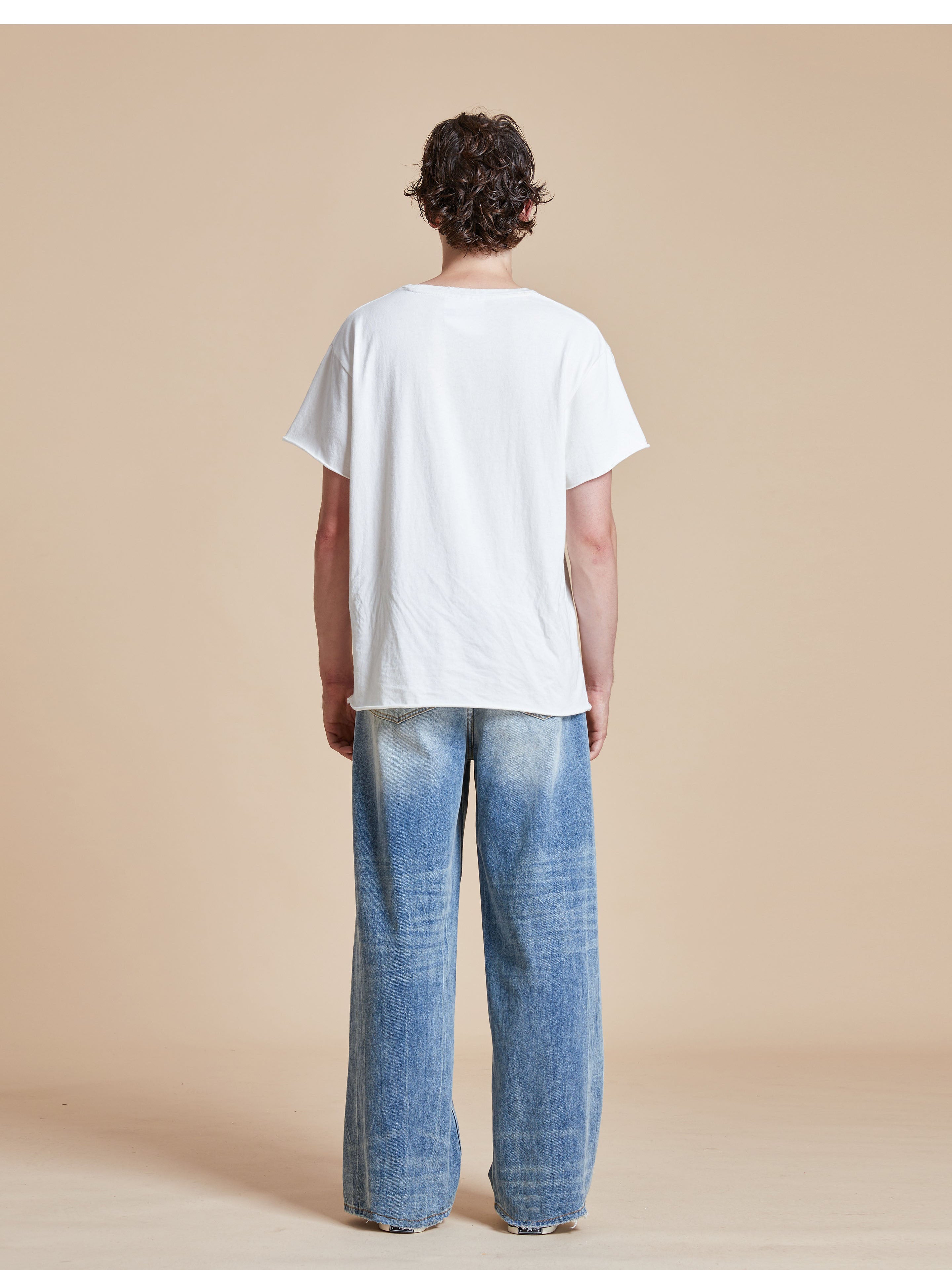 A person with curly hair stands facing away, wearing a white t-shirt and Found's Lacy Baggy Jeans featuring vintage blue gradient wash patterns and distressed hems, against a beige background.