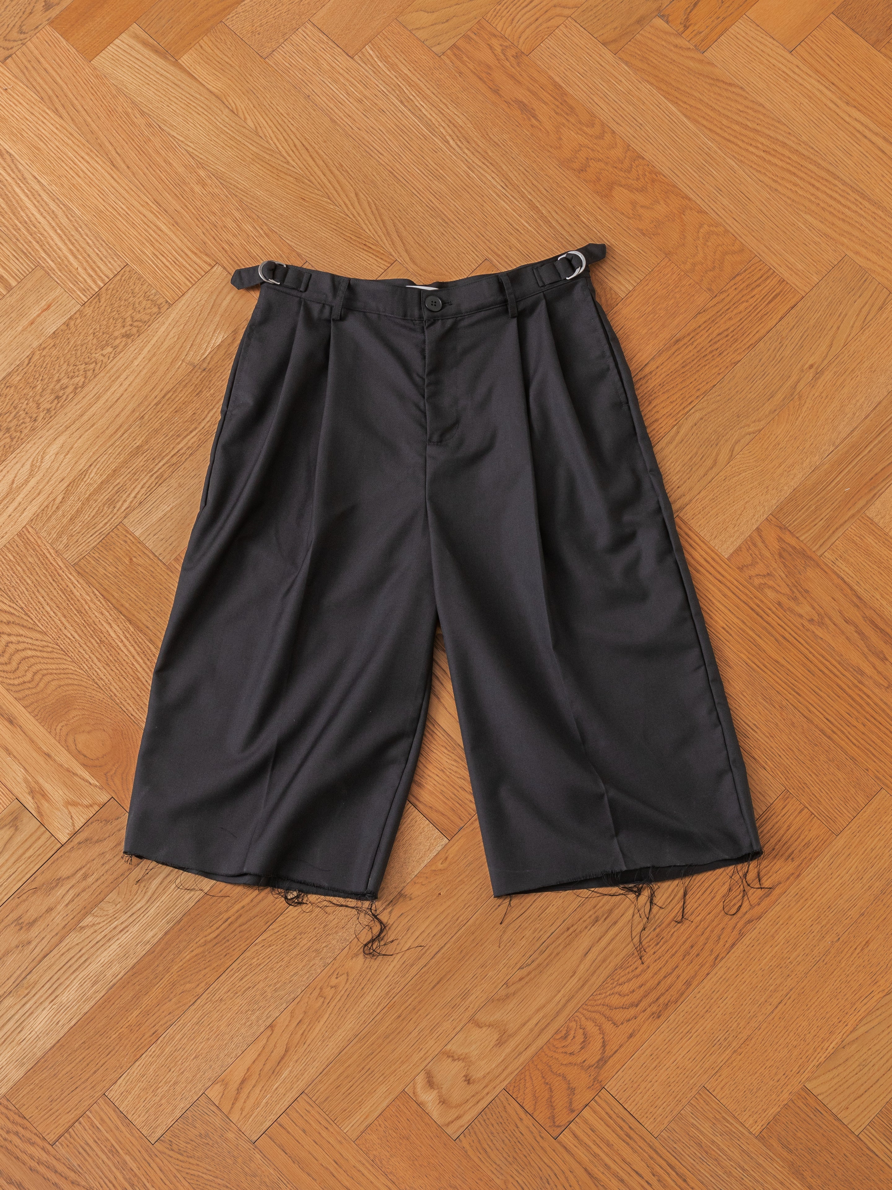 The FOUND Pleated Trouser Shorts, featuring knee-length design with a button closure, belt loops, and frayed hems, are laid flat on a wooden herringbone-patterned floor.