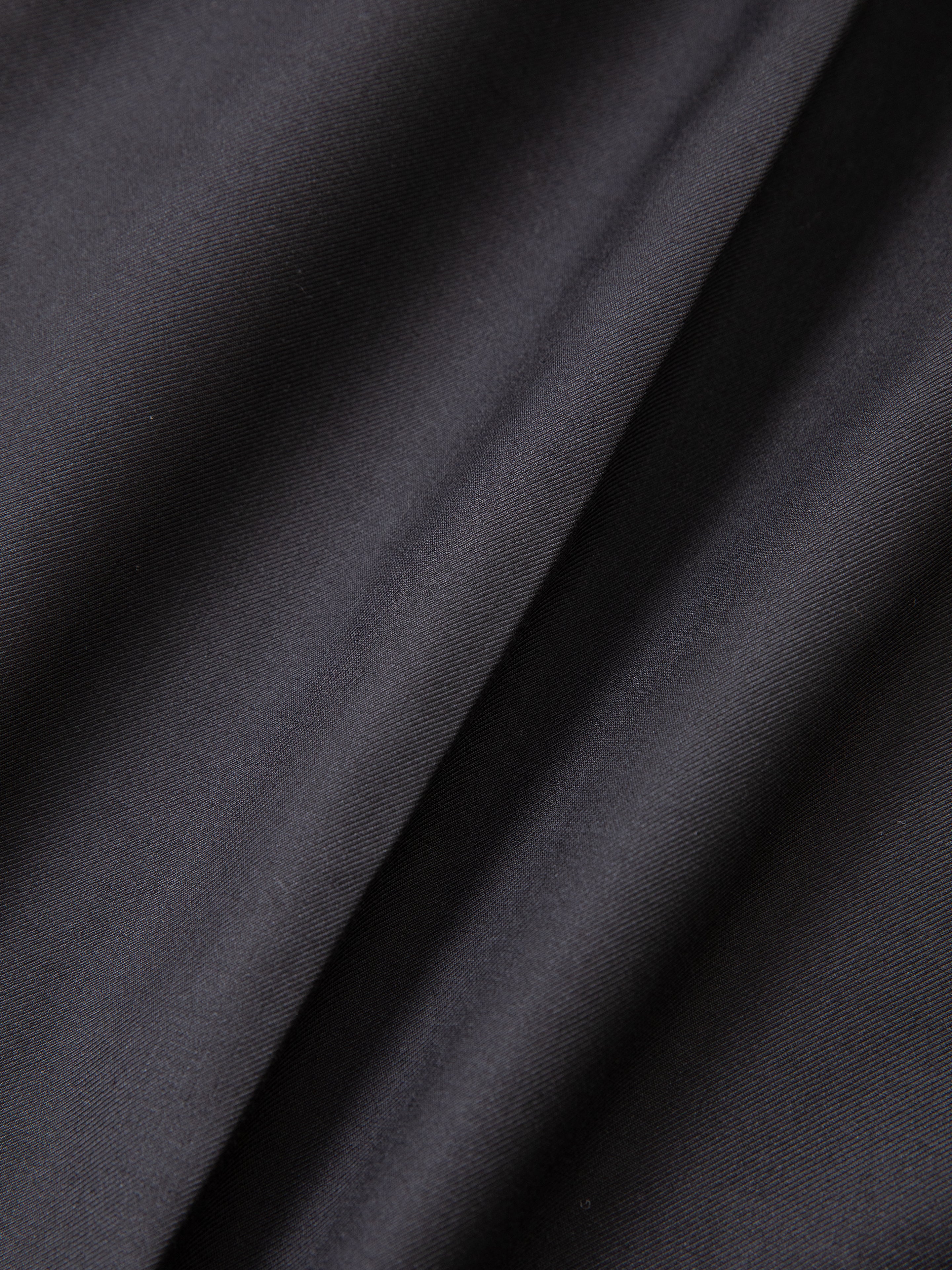 Close-up of a dark, smooth fabric with gentle folds, showcasing the double-pleated detail and adjustable waist of the Pleated Trouser Shorts by FOUND.