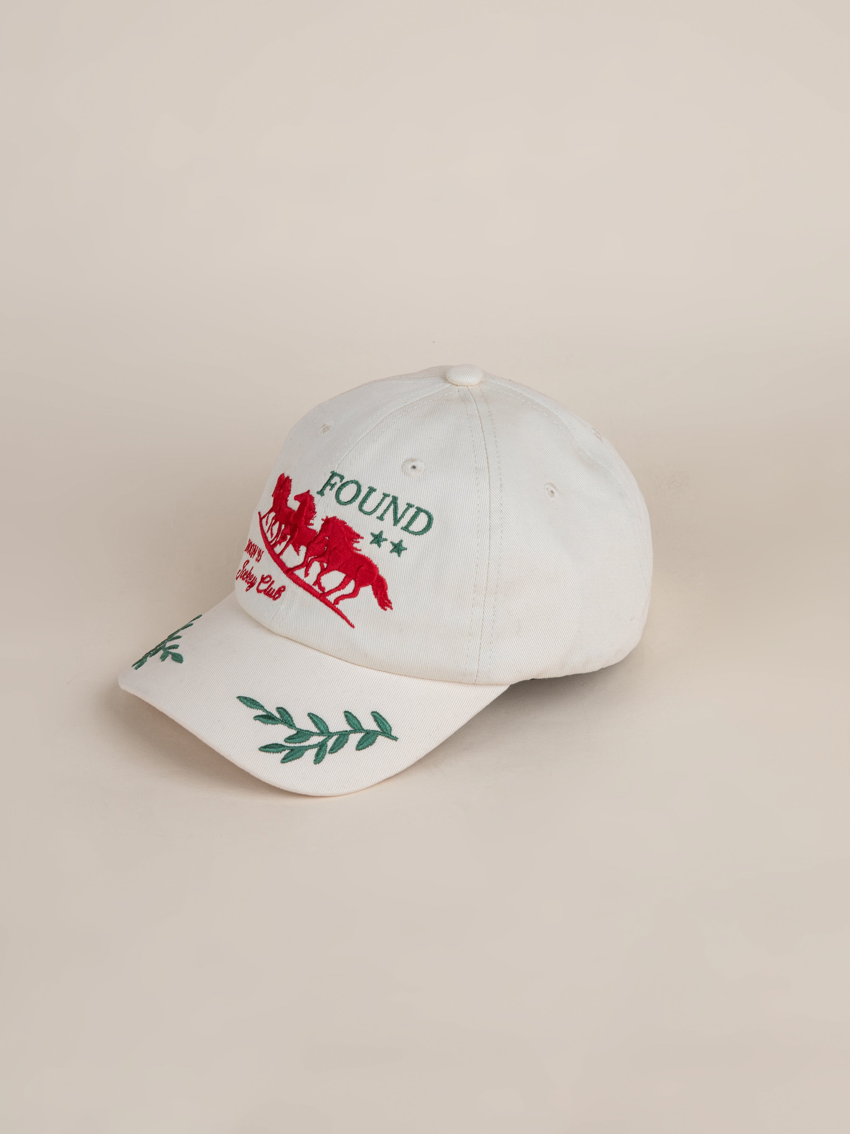 Introducing the Jockey Club Found Cap by FOUND: a stylish white cap adorned with intricate red embroidery of horses and the FOUND logo, complemented by green leaf designs on the brim.