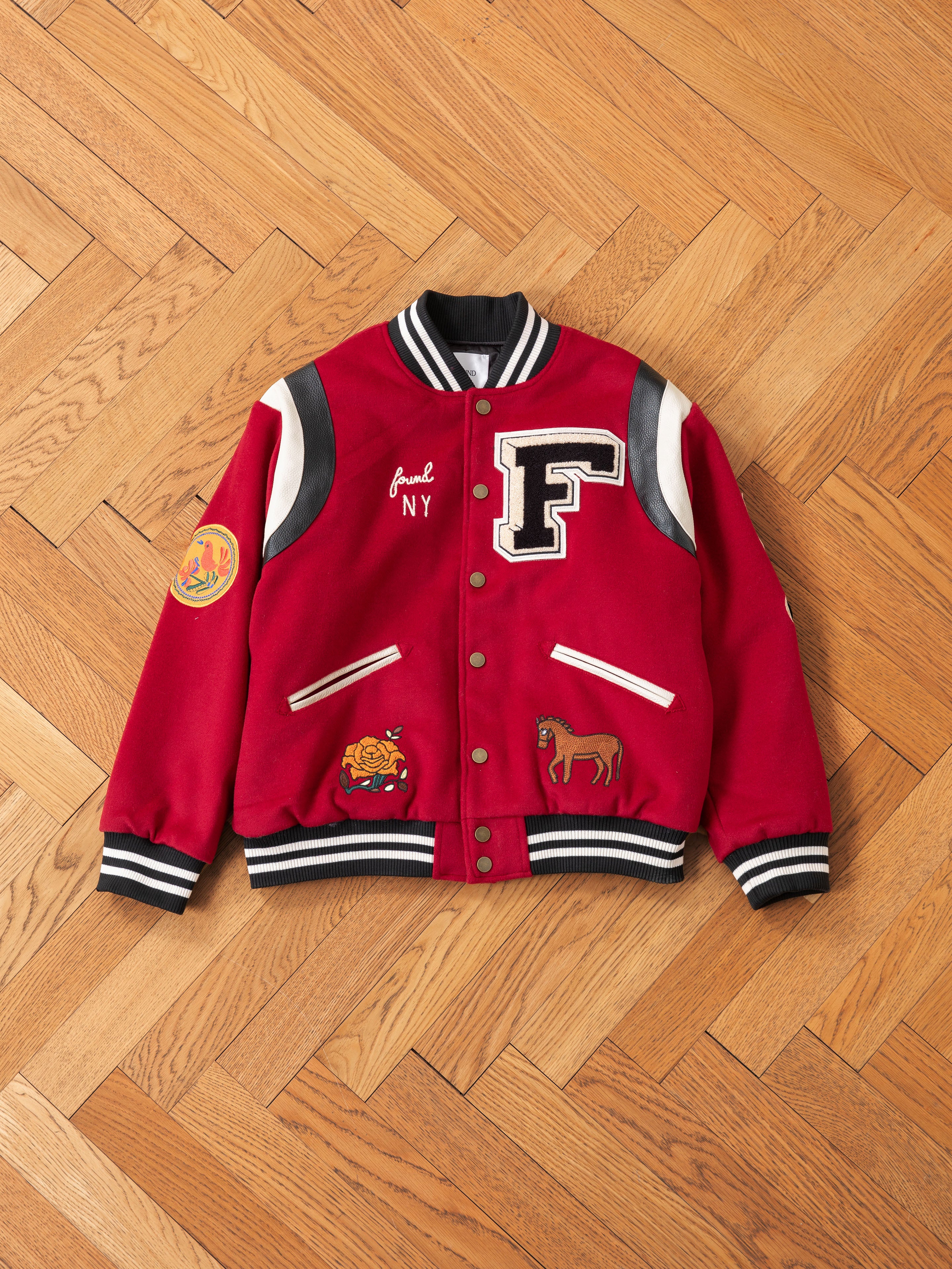 A FOUND red Letterman Varsity Jacket with black and white accents, a chenille chest logo, and patches on the front and sleeves is displayed on a wooden herringbone floor.