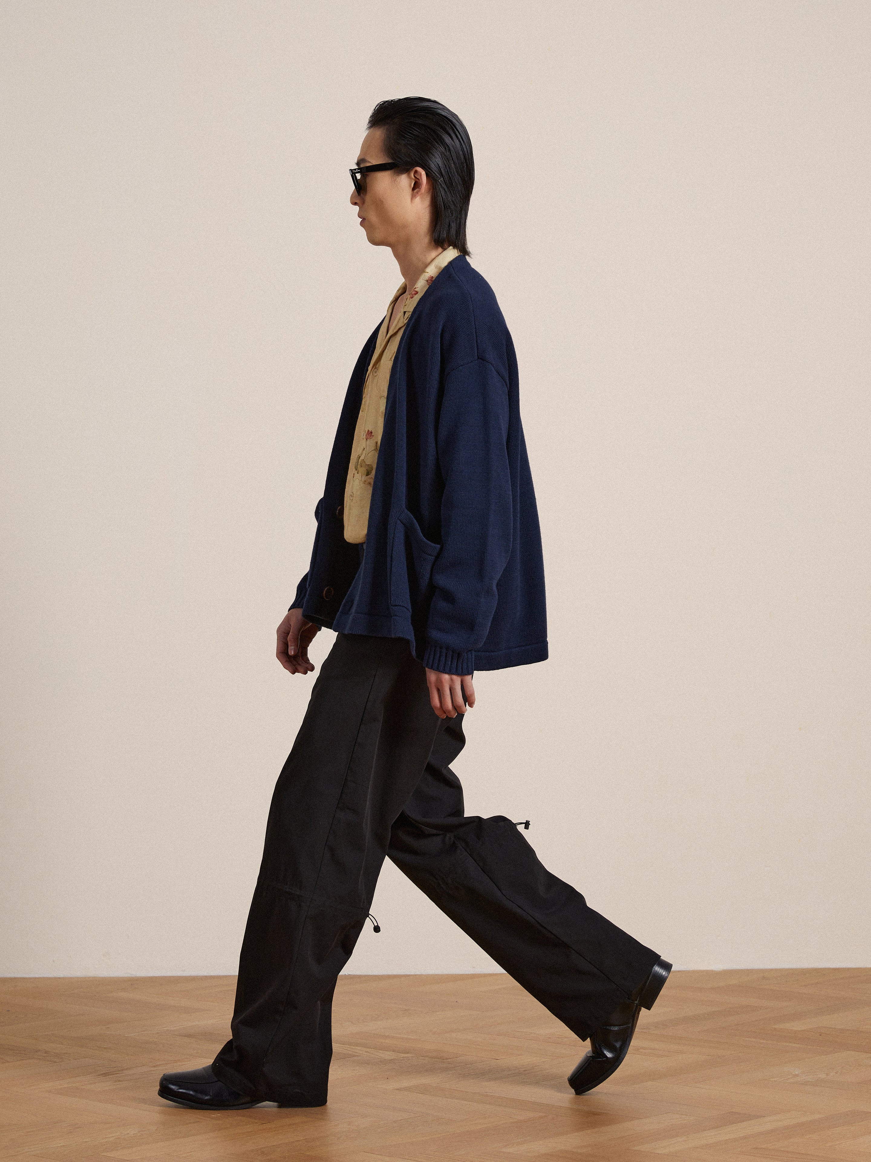 A man in sunglasses, wearing a navy jacket, beige shirt, and black Found Tencel pleated pants, walks across a beige background.