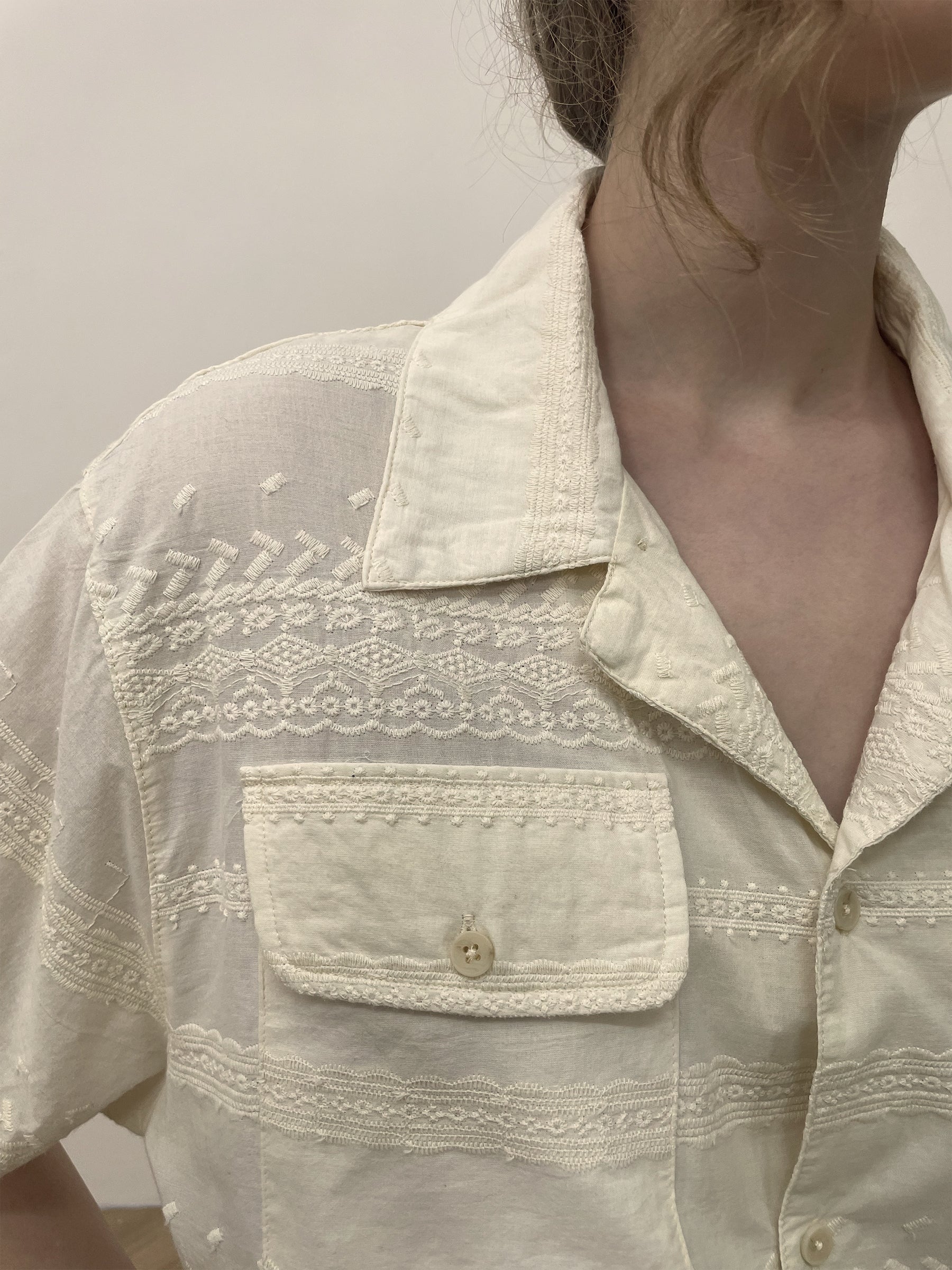 Close-up of a person wearing an off-white Lace SS Camp Shirt by Found, featuring intricate lace detailing and embroidery on the front. The premium cotton fabric enhances the elegance of the short-sleeve, button-up design, showcasing the shoulder and upper torso area.