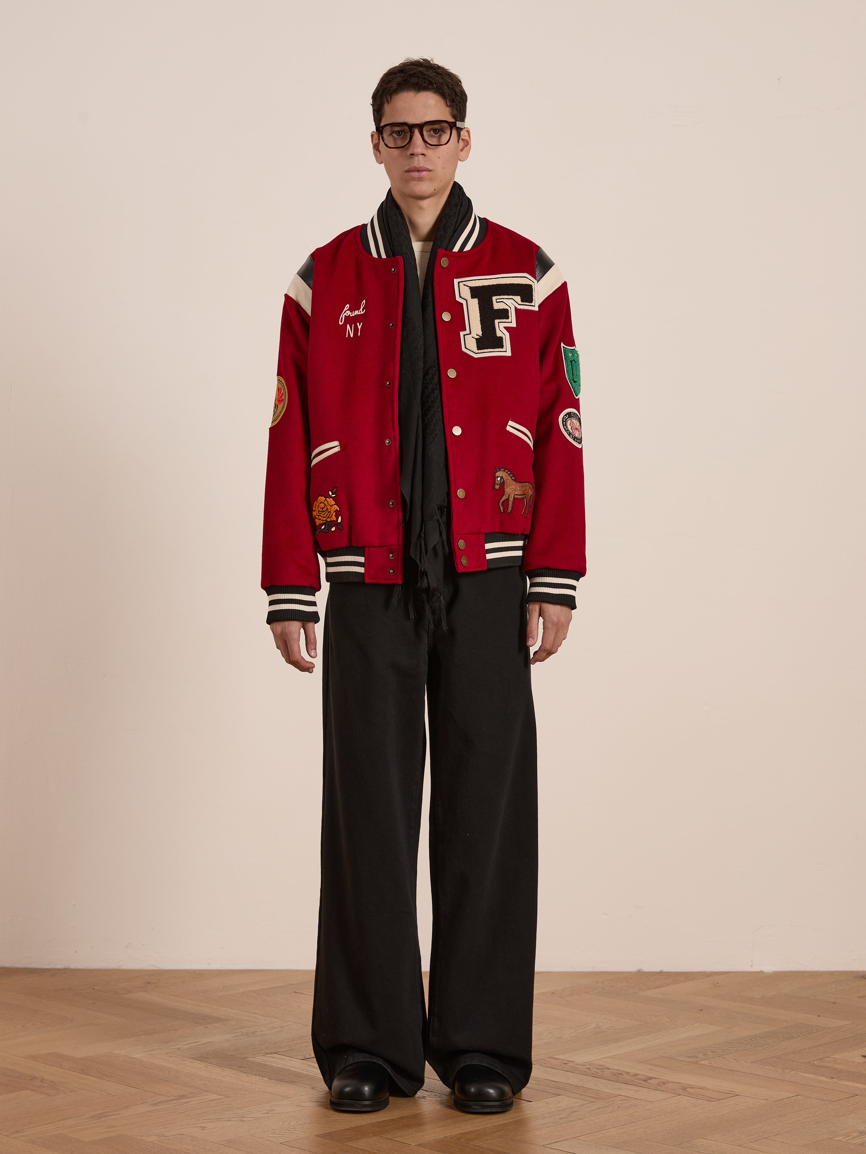 A person in a FOUND red Letterman Varsity Jacket, featuring various patches and a bold chenille chest logo, pairs it with black wide-leg pants and shoes. They stand confidently on a wooden floor against a plain wall, embodying style from ethically sourced materials.