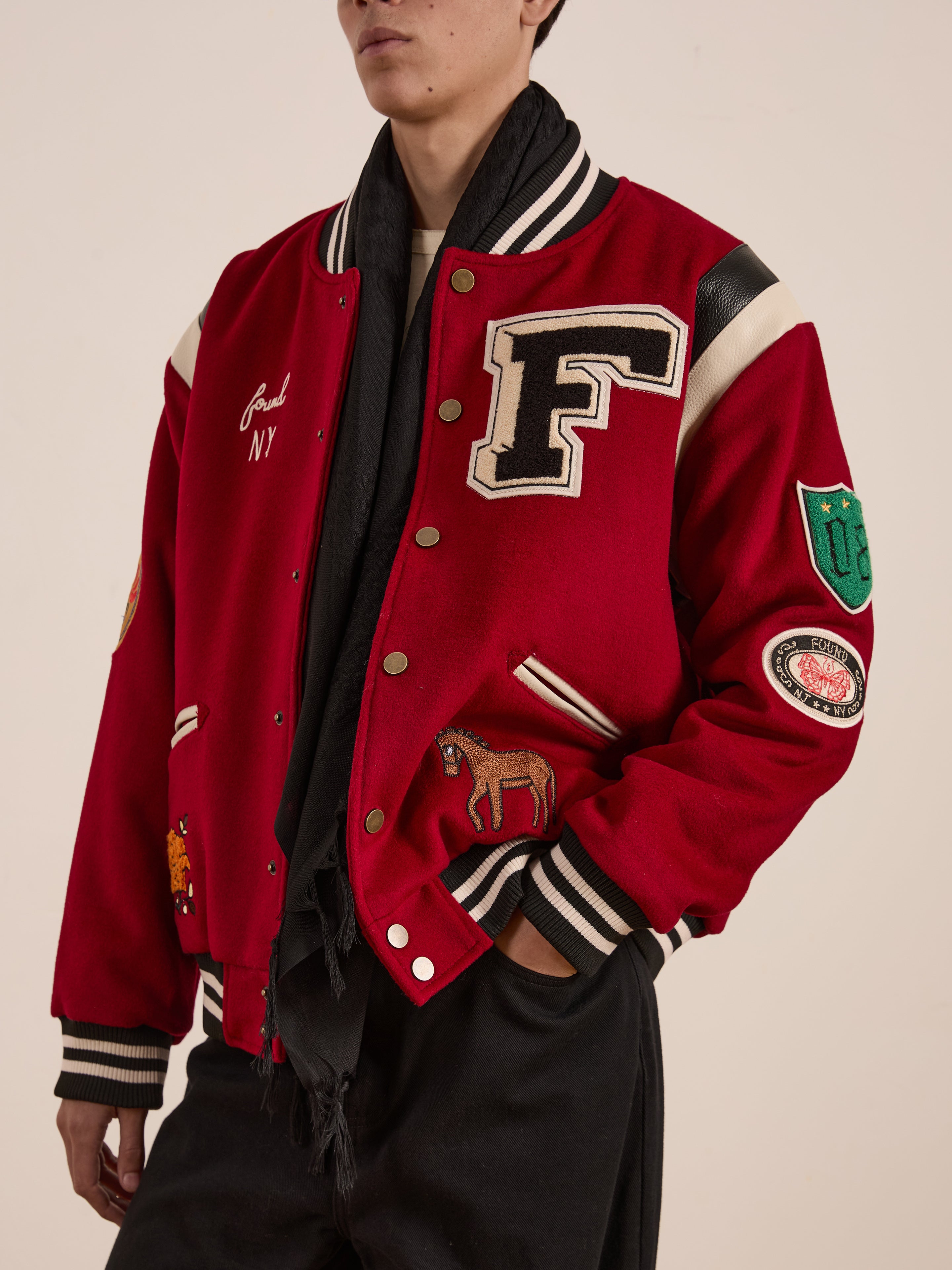 A person wearing FOUND's Letterman Varsity Jacket featuring a large chenille "F," horse, and team logos in various patches, paired with a black scarf and ethically sourced pants.