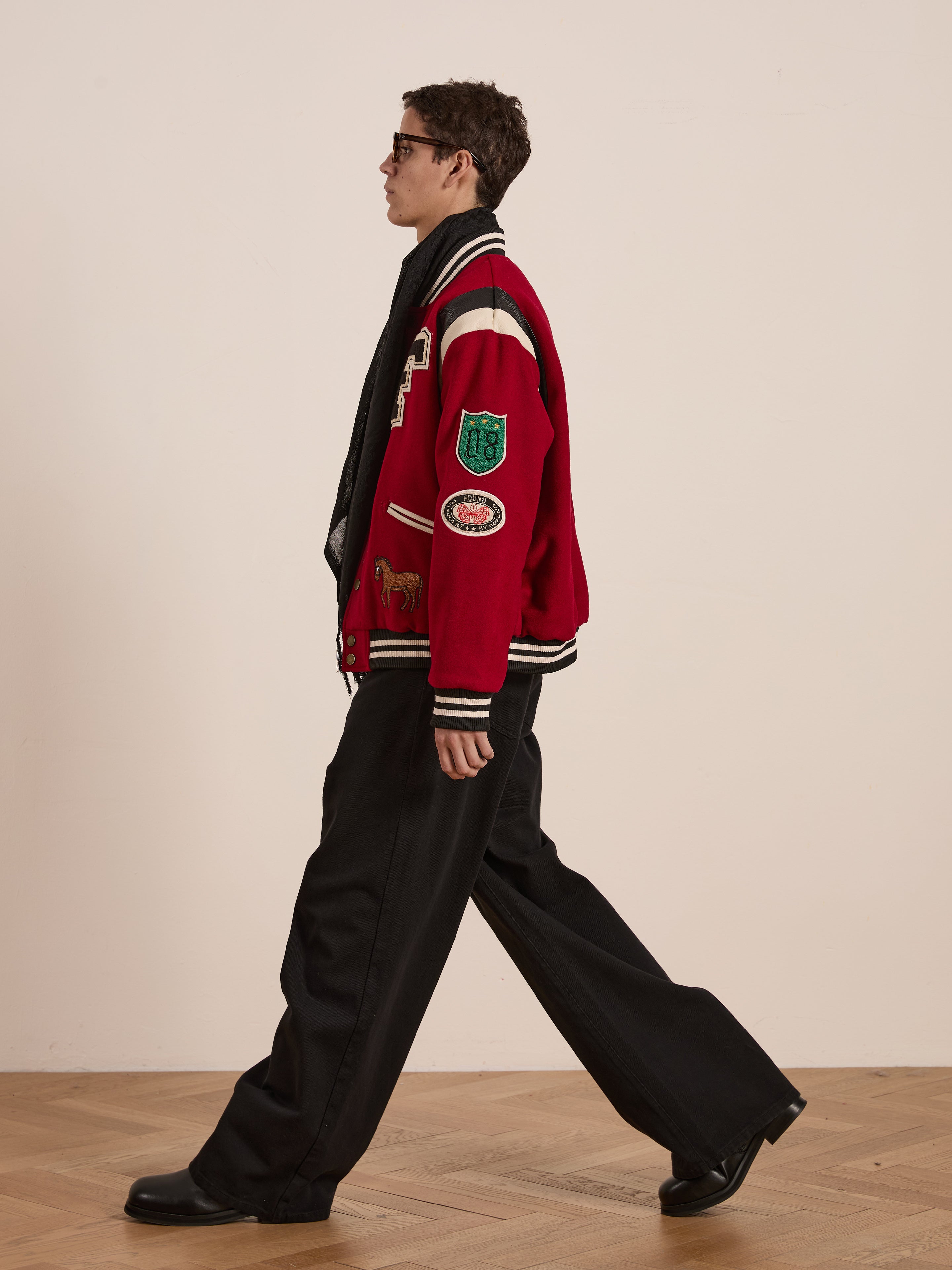 A person in FOUND's Letterman Varsity Jacket with a red chenille logo, paired with black pants and glasses, strolls across a wooden floor. The unisex relaxed fit adds to the effortless style against the plain backdrop.