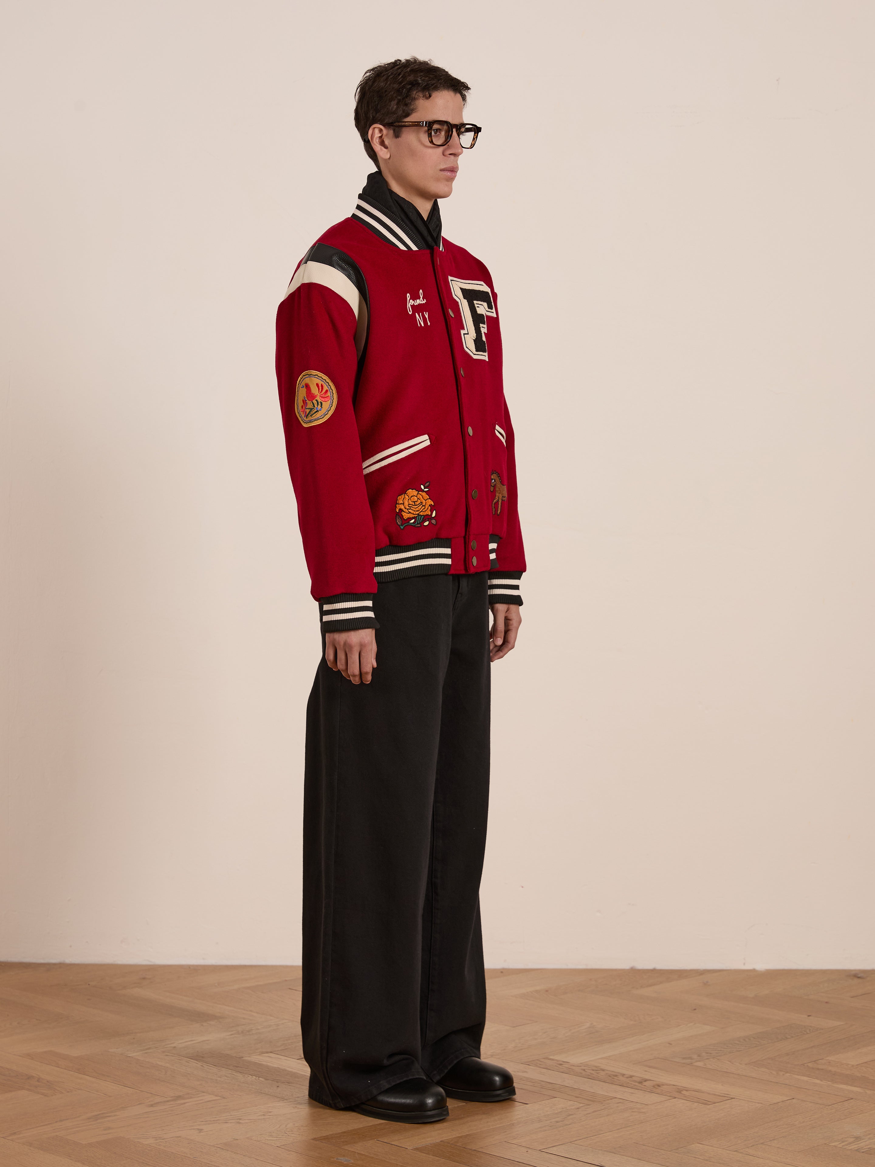 An individual poses against a plain wall wearing FOUND's red Letterman Varsity Jacket, featuring vibrant patches and a Chenille chest logo. They pair it with unisex relaxed-fit wide black pants on a wooden floor.