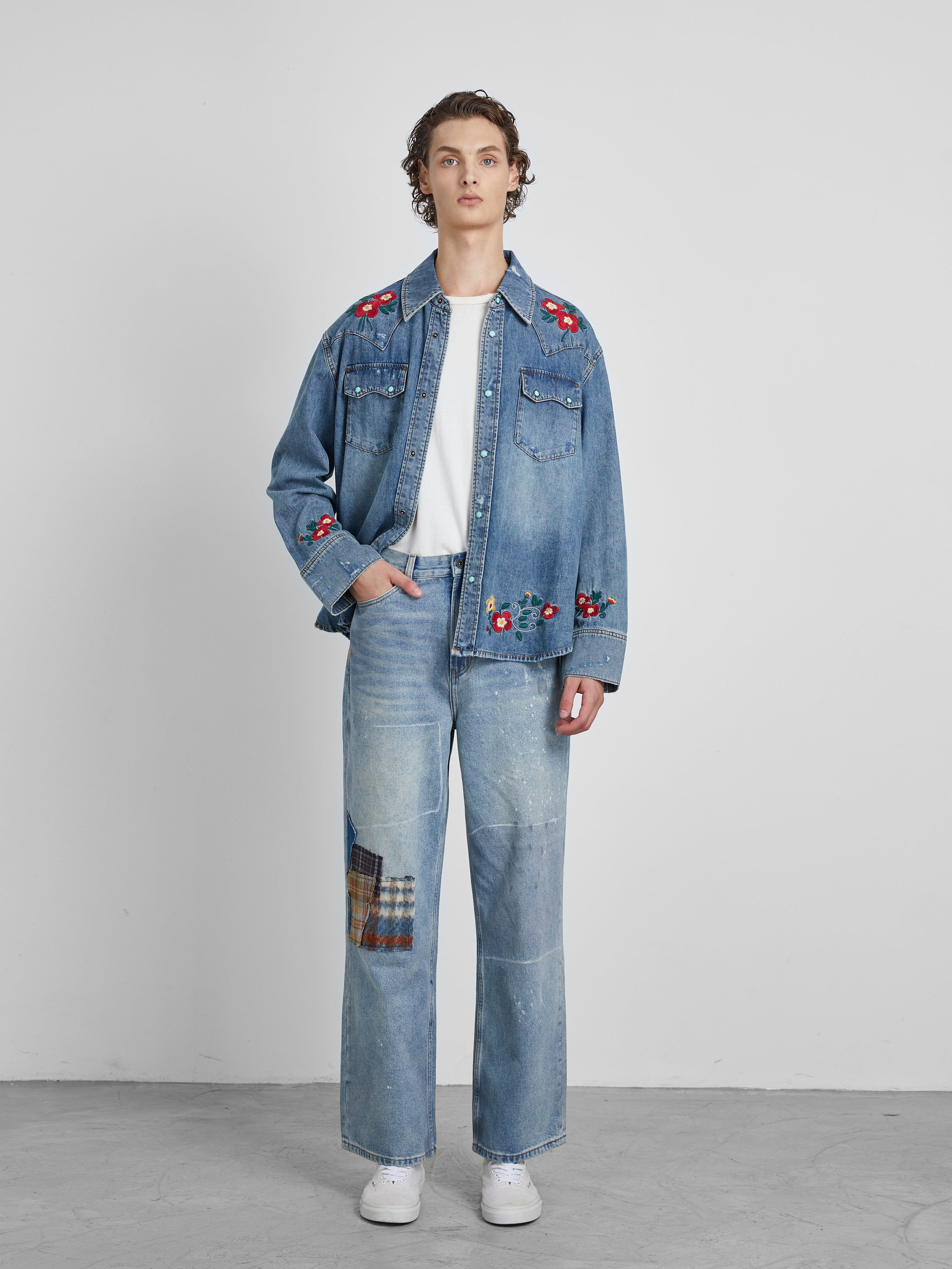 A person dressed in Profound's Mix Patch Jeans, paired with a denim cotton jacket adorned with floral embroidery, stands against a plain backdrop.