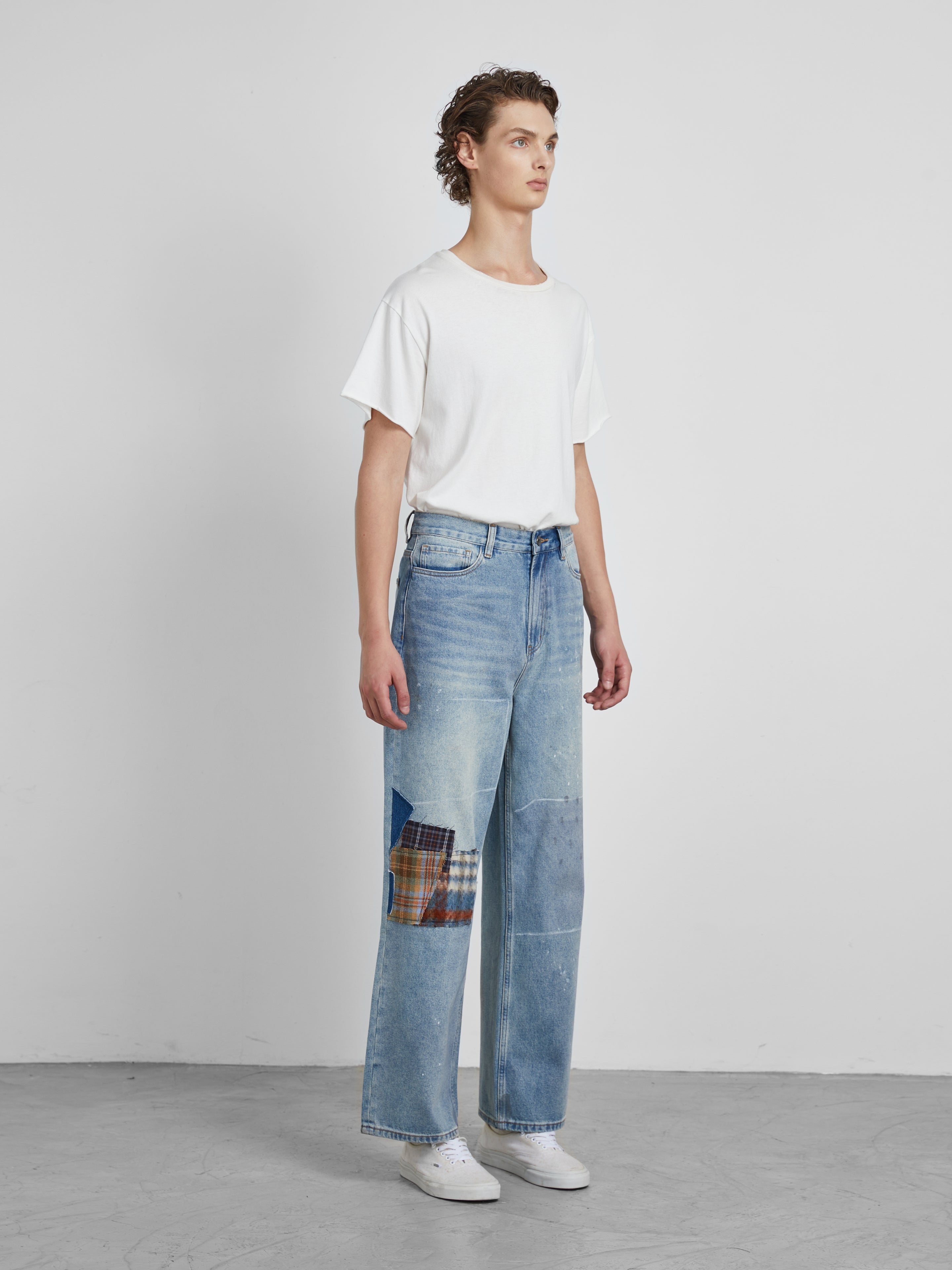 A person in a white t-shirt and Mix Patch Jeans by Profound stands against a plain white background, showcasing the intricate details of their wide-leg denim cotton pants.