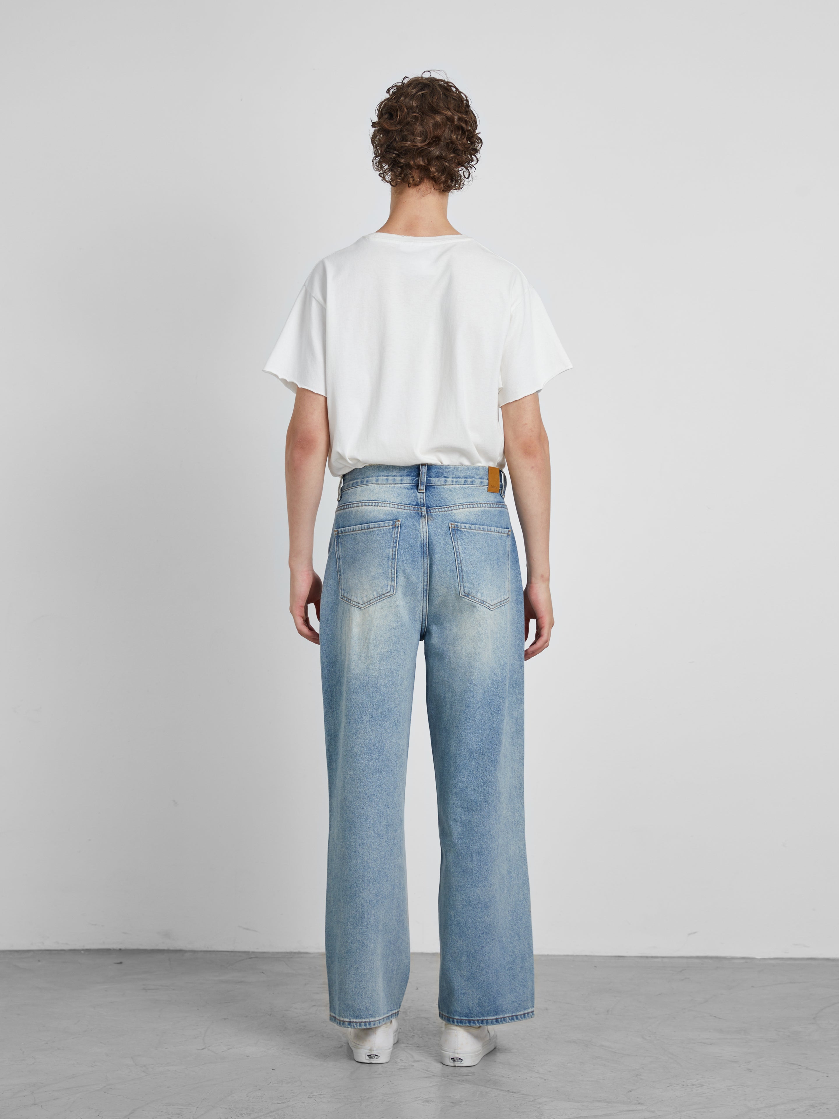 A person wearing a white t-shirt and Profound's Mix Patch Jeans stands facing a plain white wall.