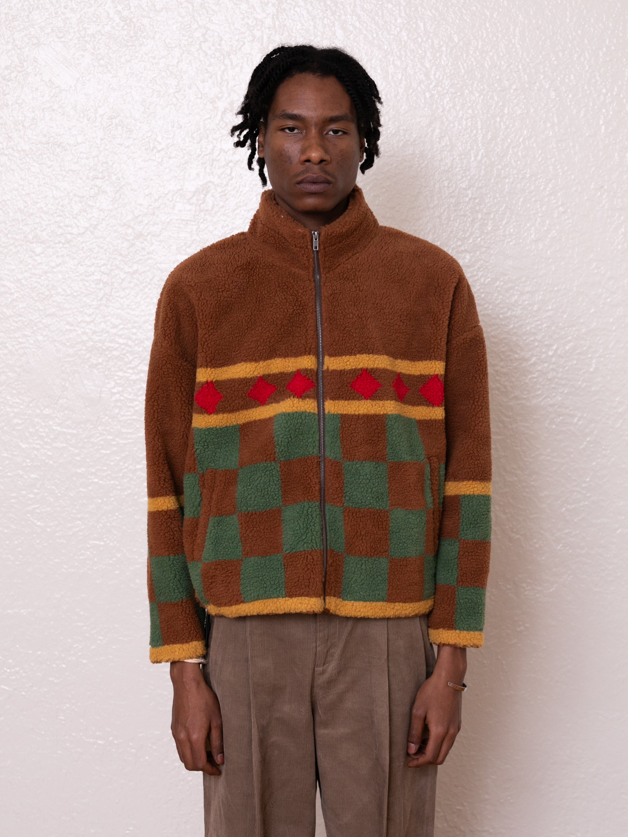 A person stands against a white textured wall, wearing FOUND's Checkered Zip-Up Sherpa in brown fleece, featuring red, green, and yellow jacquard checkered designs. Tan pants complete the ensemble, while an antique silver YKK zipper adds a touch of elegance to the look.