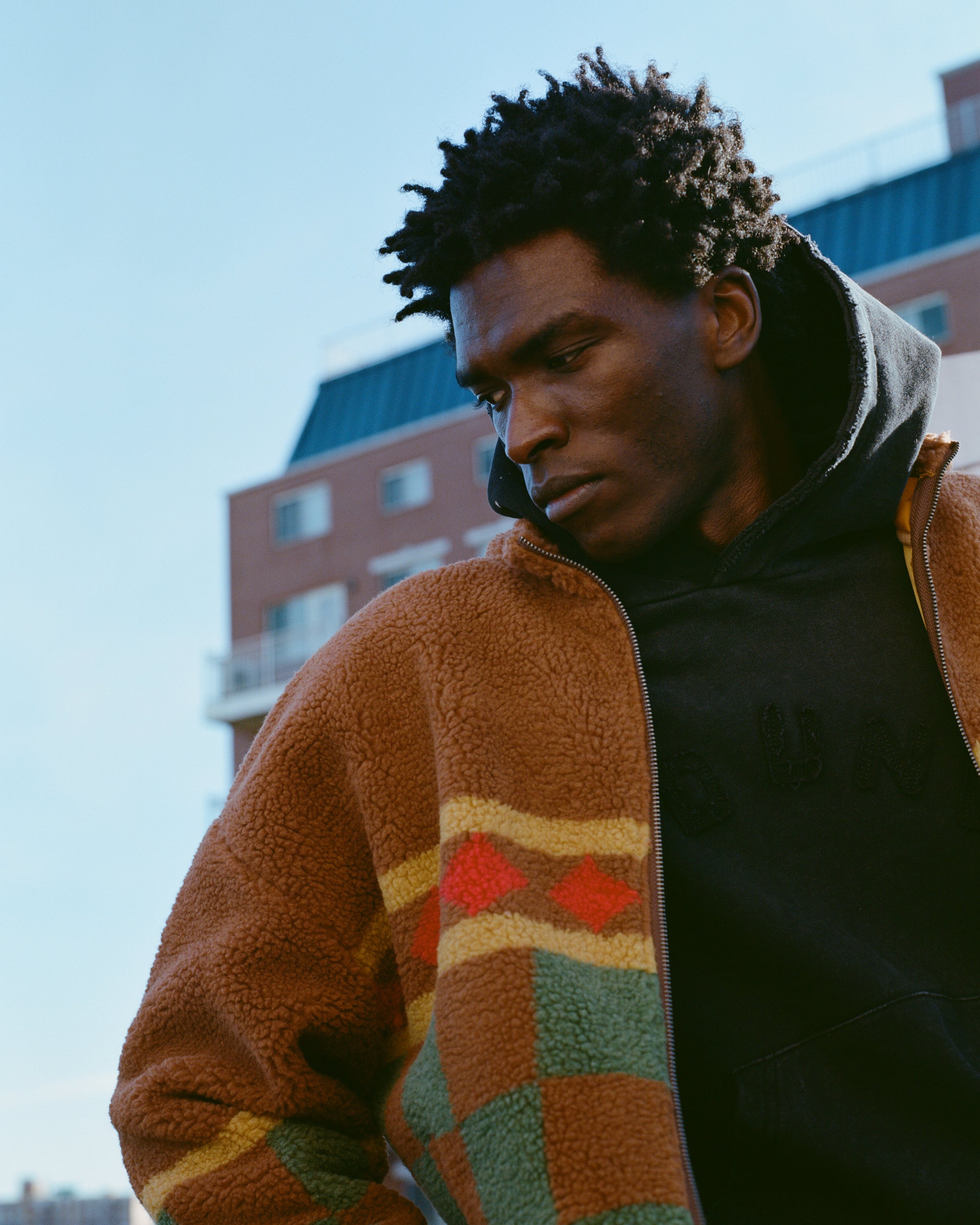 A person in a fleece jacket and hoodie stands outdoors with buildings in the background, their style enhanced by the Checkered Zip-Up Sherpa from FOUND, featuring adjustable toggles.