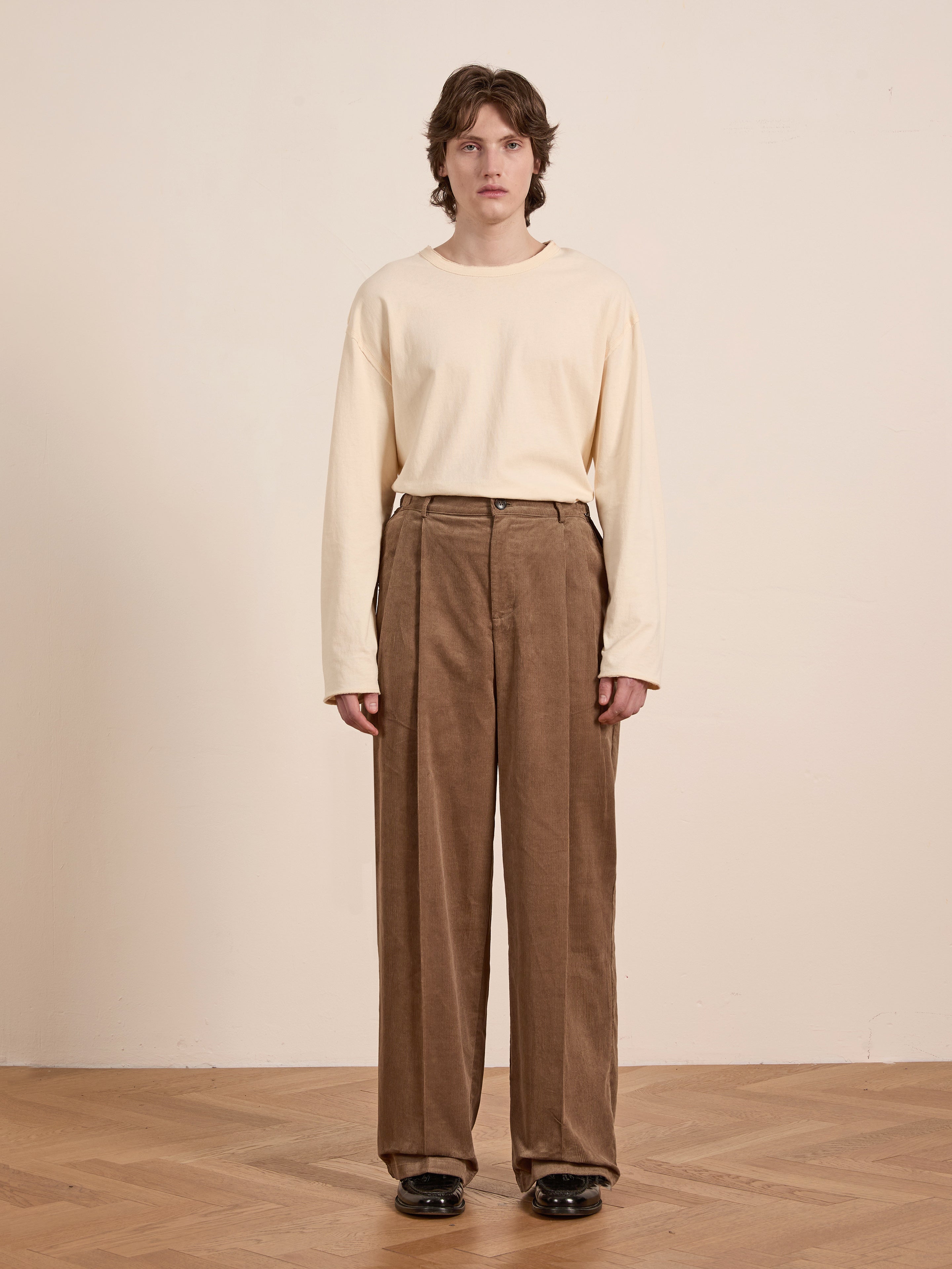 An individual in a cream long-sleeve shirt and FOUND’s Corduroy Pleated Trousers stands on a wooden floor against a plain beige wall.