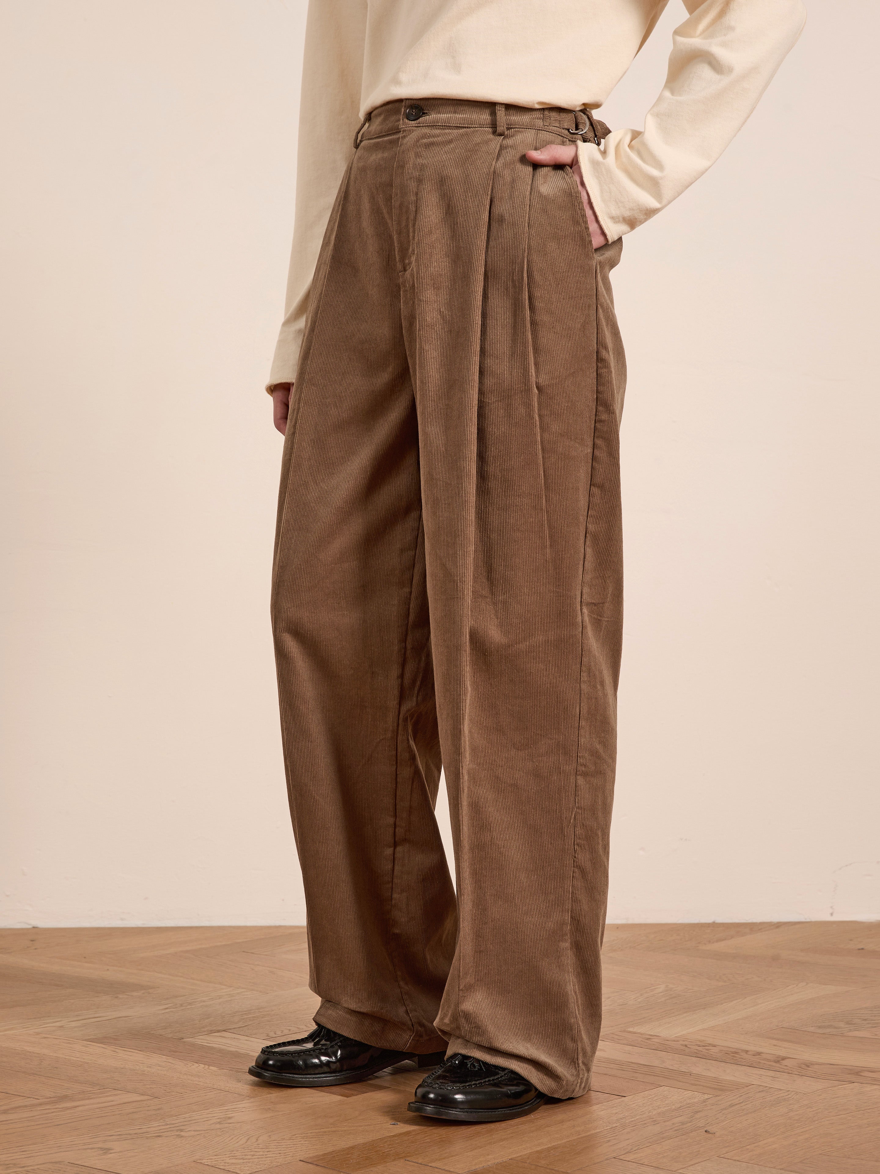 A person stands on a wooden floor wearing FOUND's unisex beige Corduroy Pleated Trousers and a cream long-sleeve shirt.