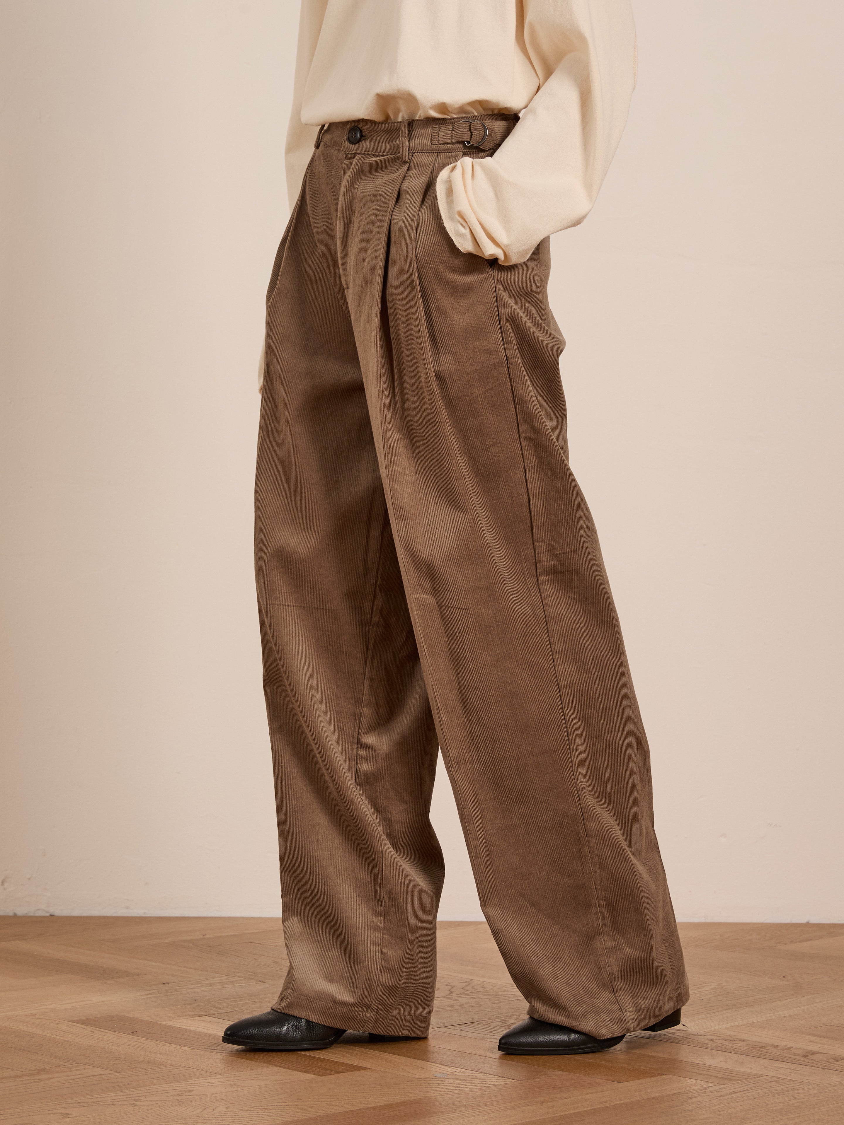 A person wearing a cream sweater, FOUND Corduroy Pleated Trousers in brown, and black shoes stands with one hand in their pocket on a wooden floor.