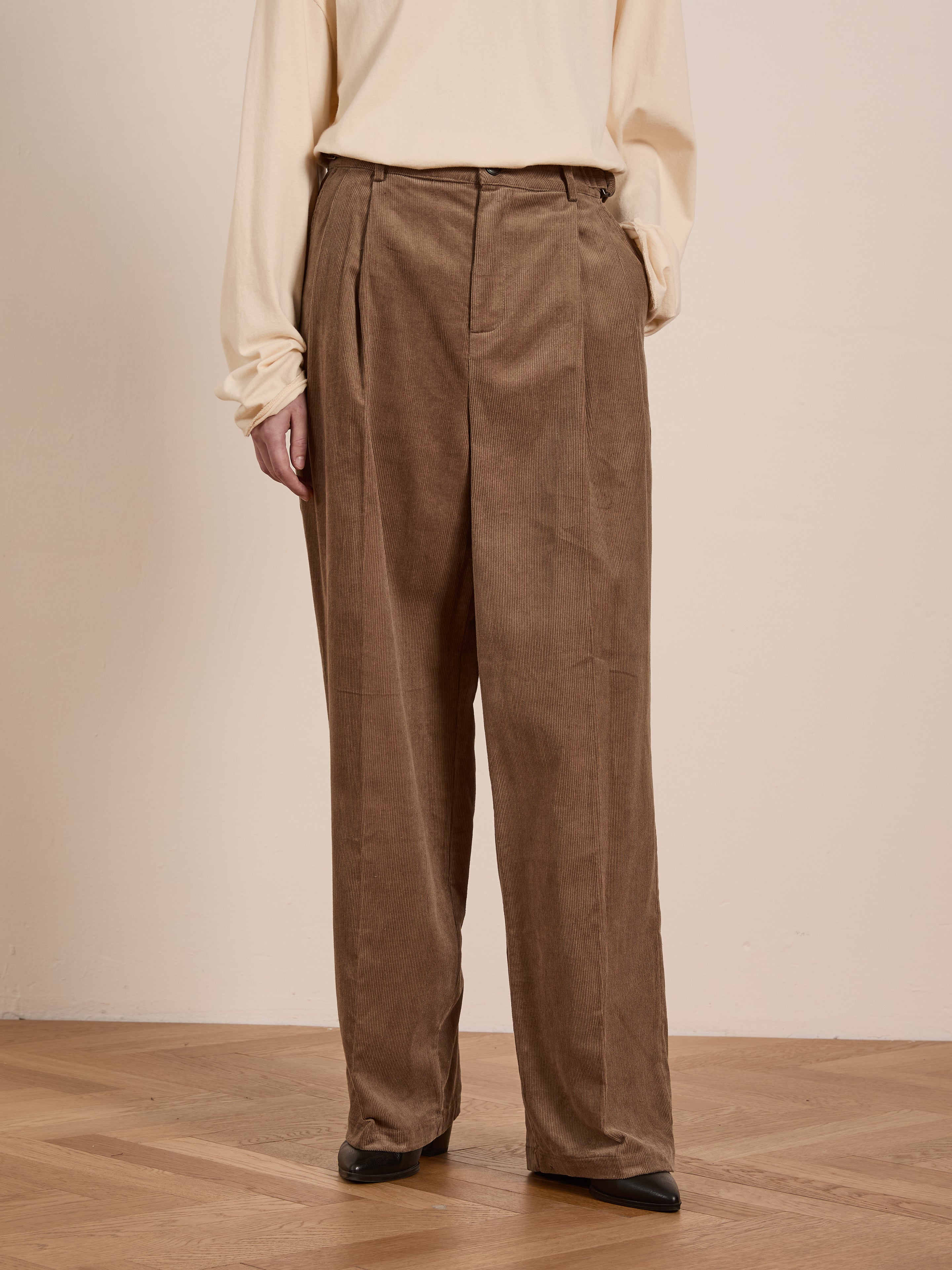 A person wearing a cream top is standing on a wooden floor, dressed in FOUND's Corduroy Pleated Trousers, featuring a loose fit and high-rise waist.