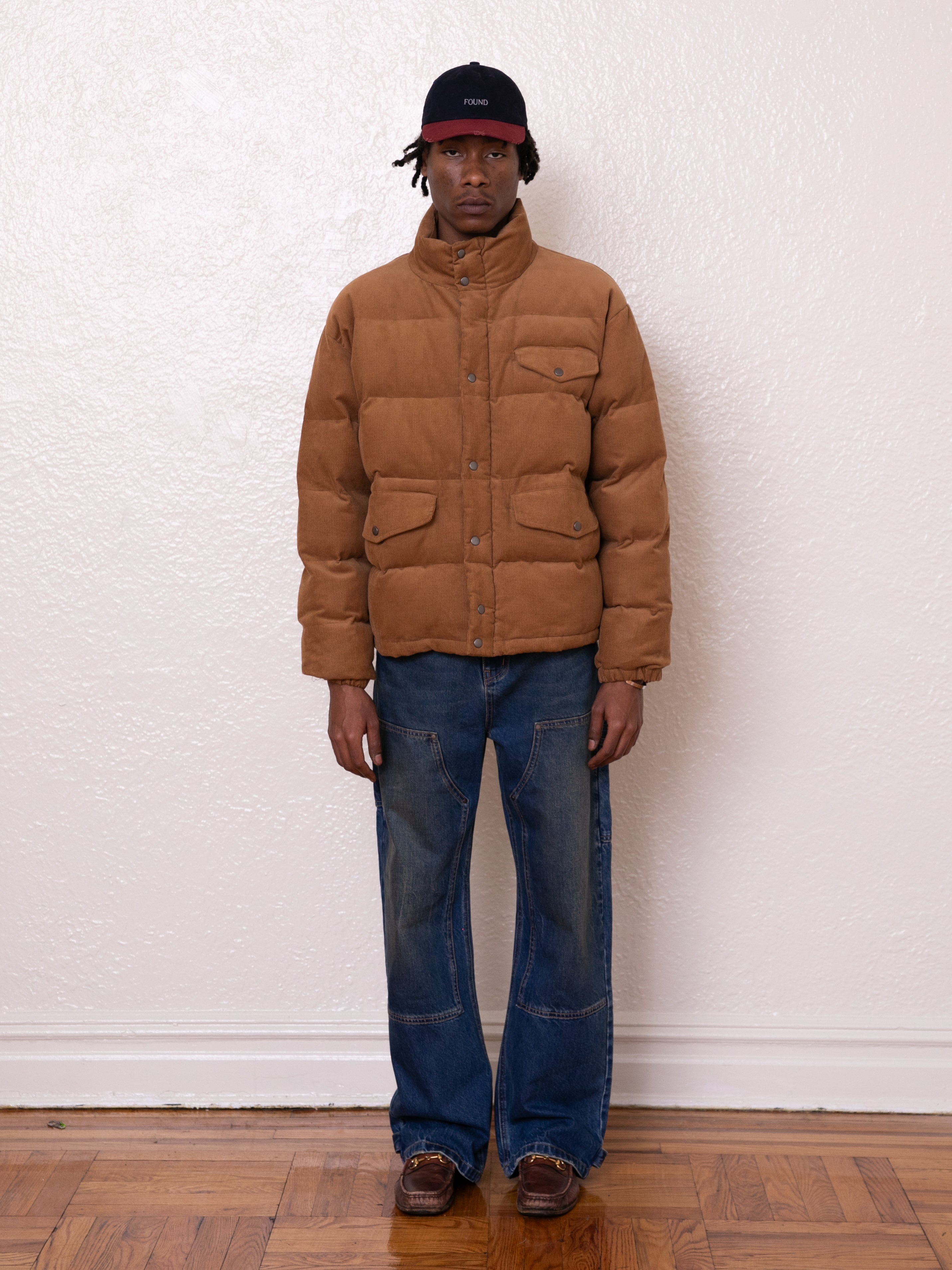 A person wearing a FOUND Corduroy Puffer Jacket in a warm tan with a relaxed boxy fit stands against a plain white wall. They are paired with blue jeans, brown shoes, and a black cap, posed comfortably on the wooden floor.