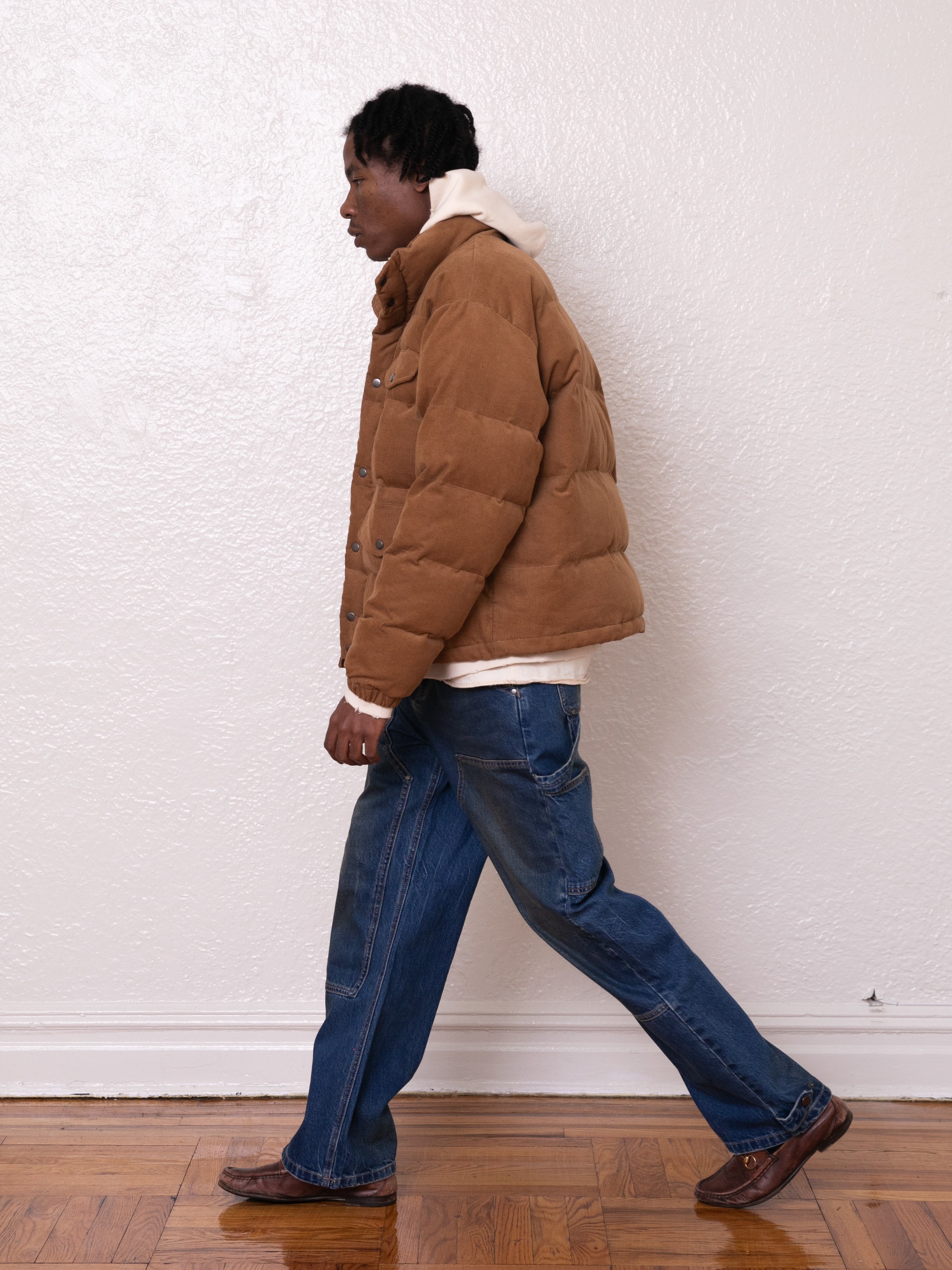 A person in a FOUND Corduroy Puffer Jacket, featuring a warm tan color and relaxed boxy fit, paired with a white hoodie and jeans, walks across a wooden floor against a white wall background.