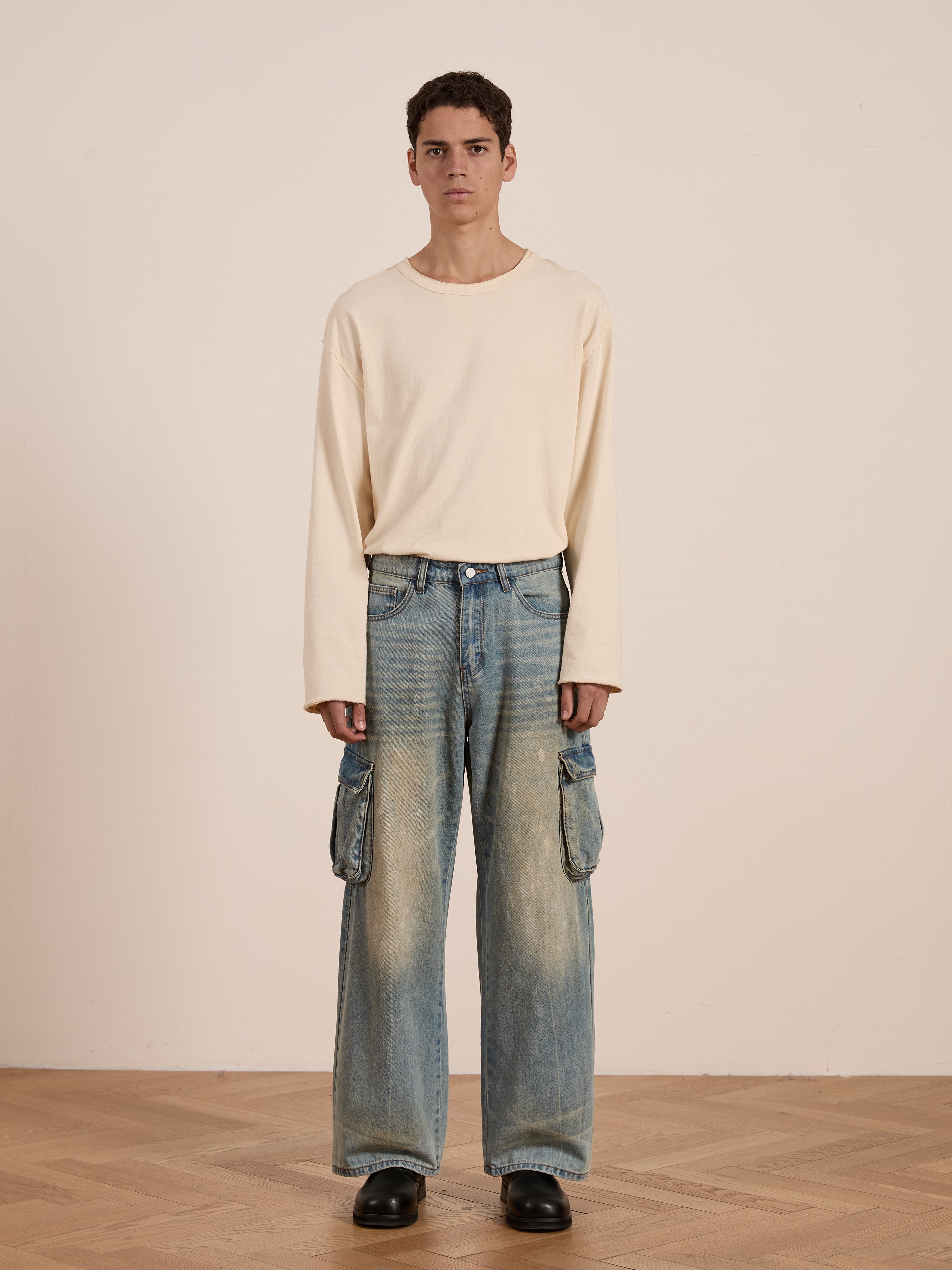 A model wearing a cream long-sleeve shirt and relaxed-fit wide-leg distressed FOUND Cargo Jeans, paired with black shoes, stands on a wooden floor against a plain beige backdrop, highlighting a subtle utilitarian edge.