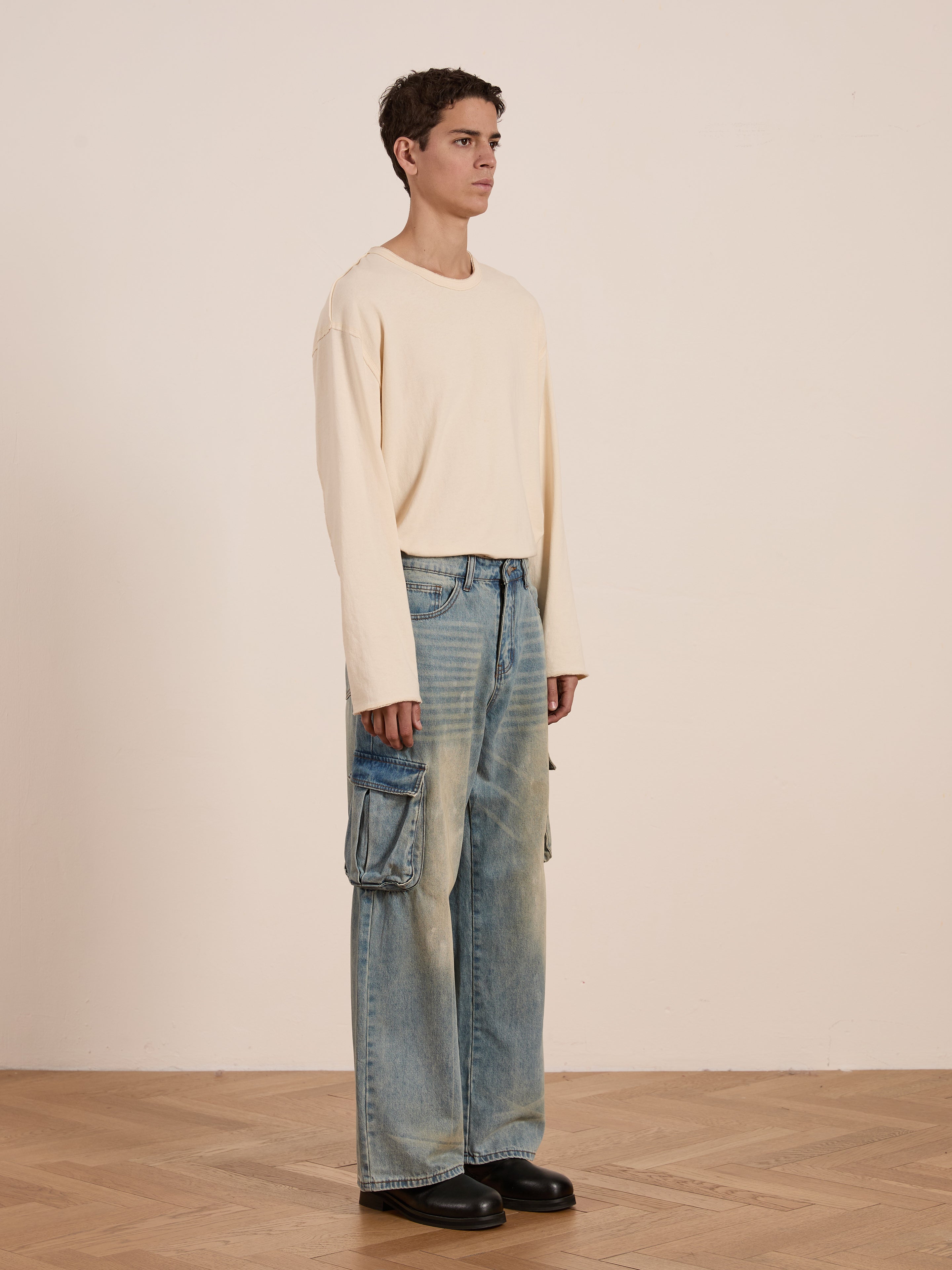 In a minimalist room with wooden floors, someone models a relaxed fit ensemble featuring FOUND Cargo Jeans in blue, paired with a beige long-sleeve shirt. Black shoes complete the utilitarian look against the plain wall.