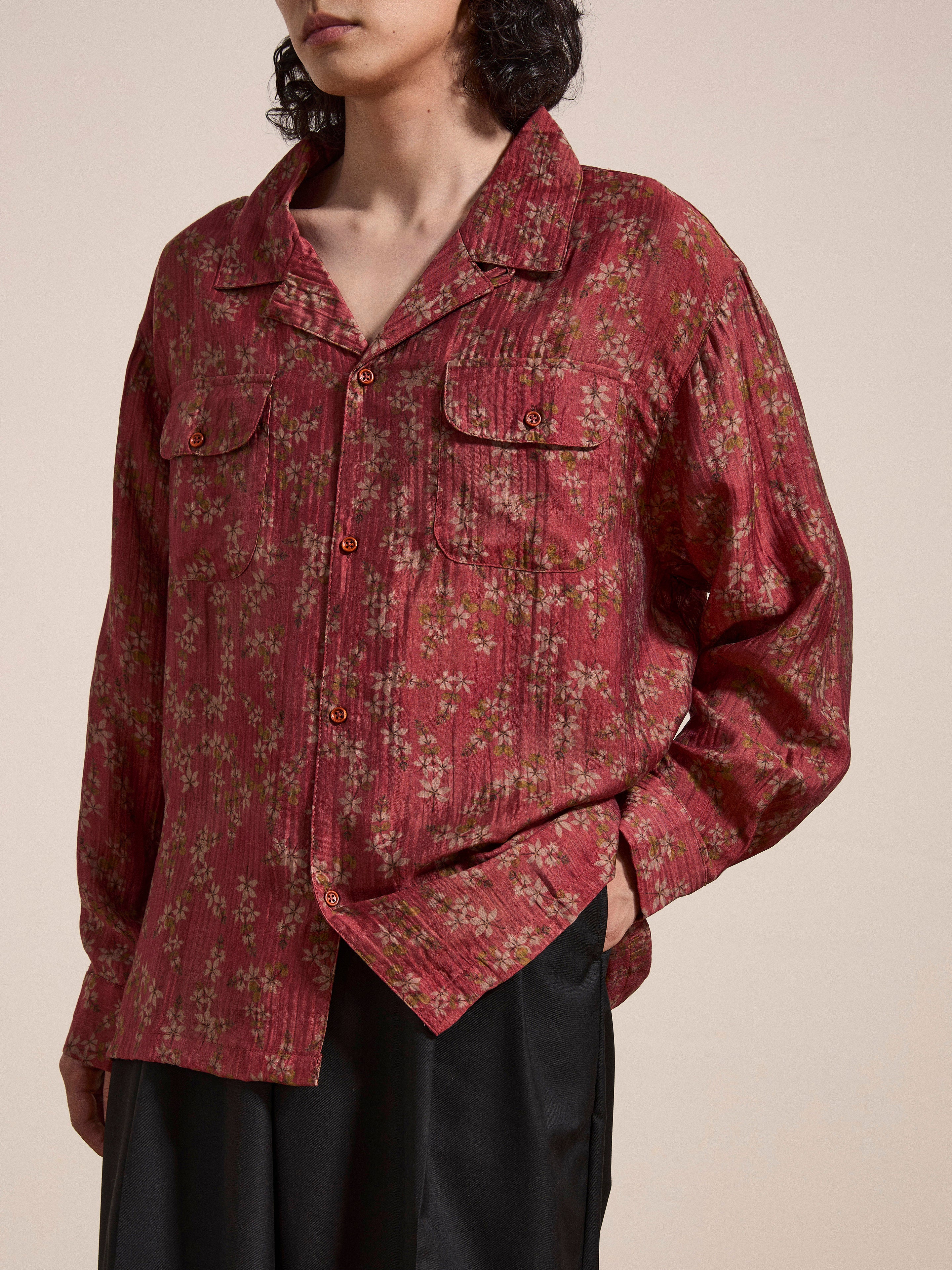 A person wearing a FOUND Charlot LS Camp Shirt, featuring a loose, long-sleeved red floral pattern and dual chest pockets, with hands partially in black pants pockets.