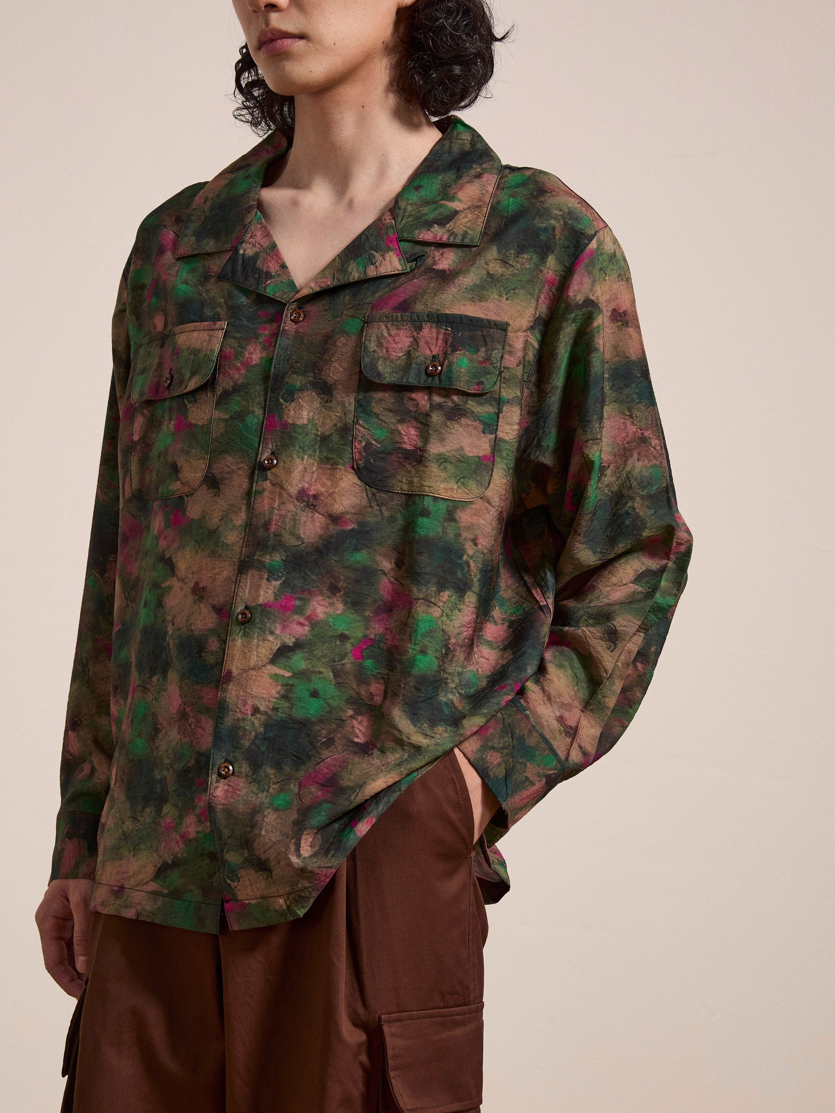 A person wearing the FOUND Charlot LS Camp Shirt, which features a green and brown camouflage pattern with dual chest pockets, stands with one hand in their pocket. They are also wearing brown pants.