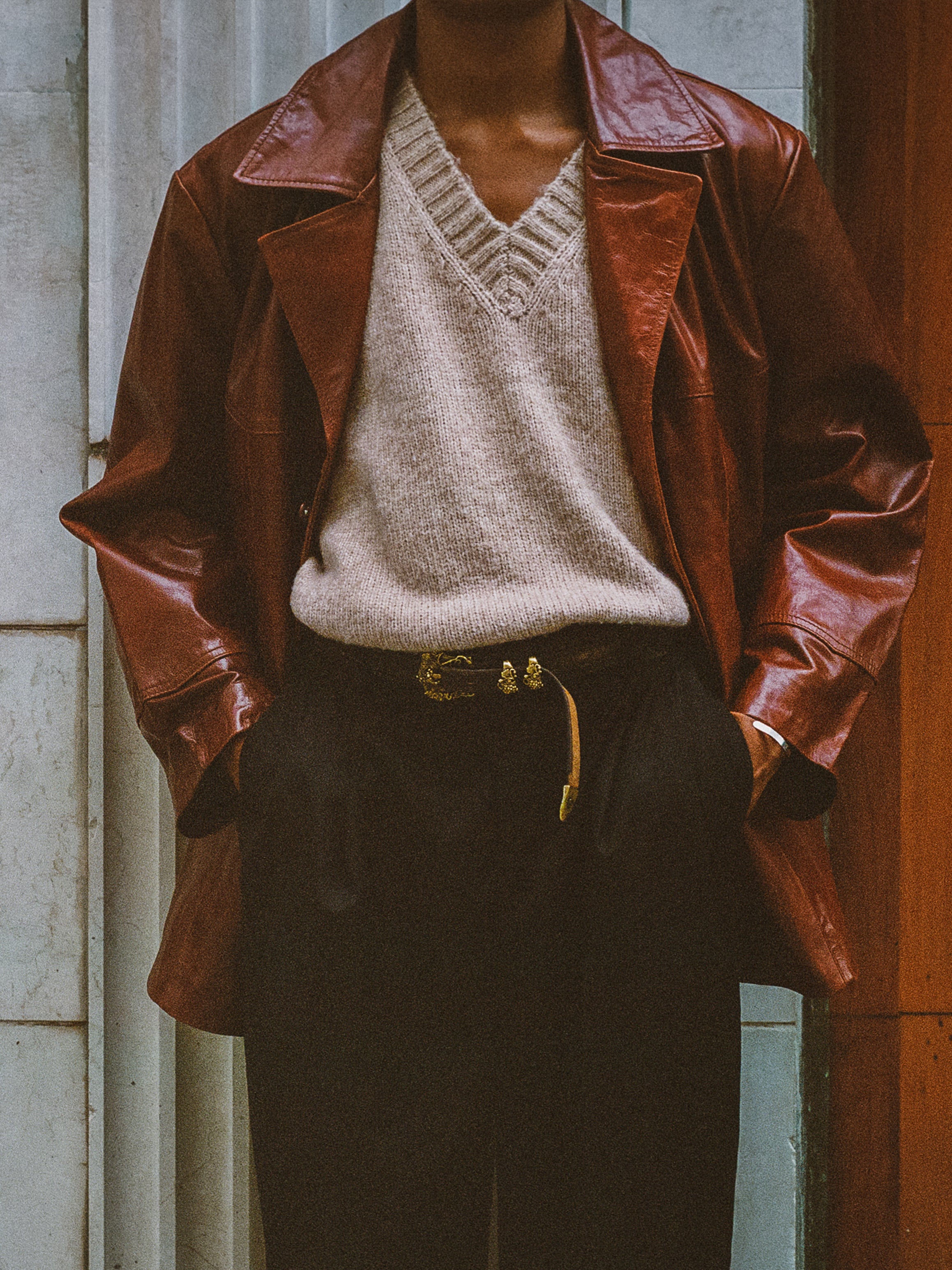 A person in a lightweight Claret Western Leather Blazer by FOUND, a beige v-neck sweater, and black pants stands against a white and wooden panel background.