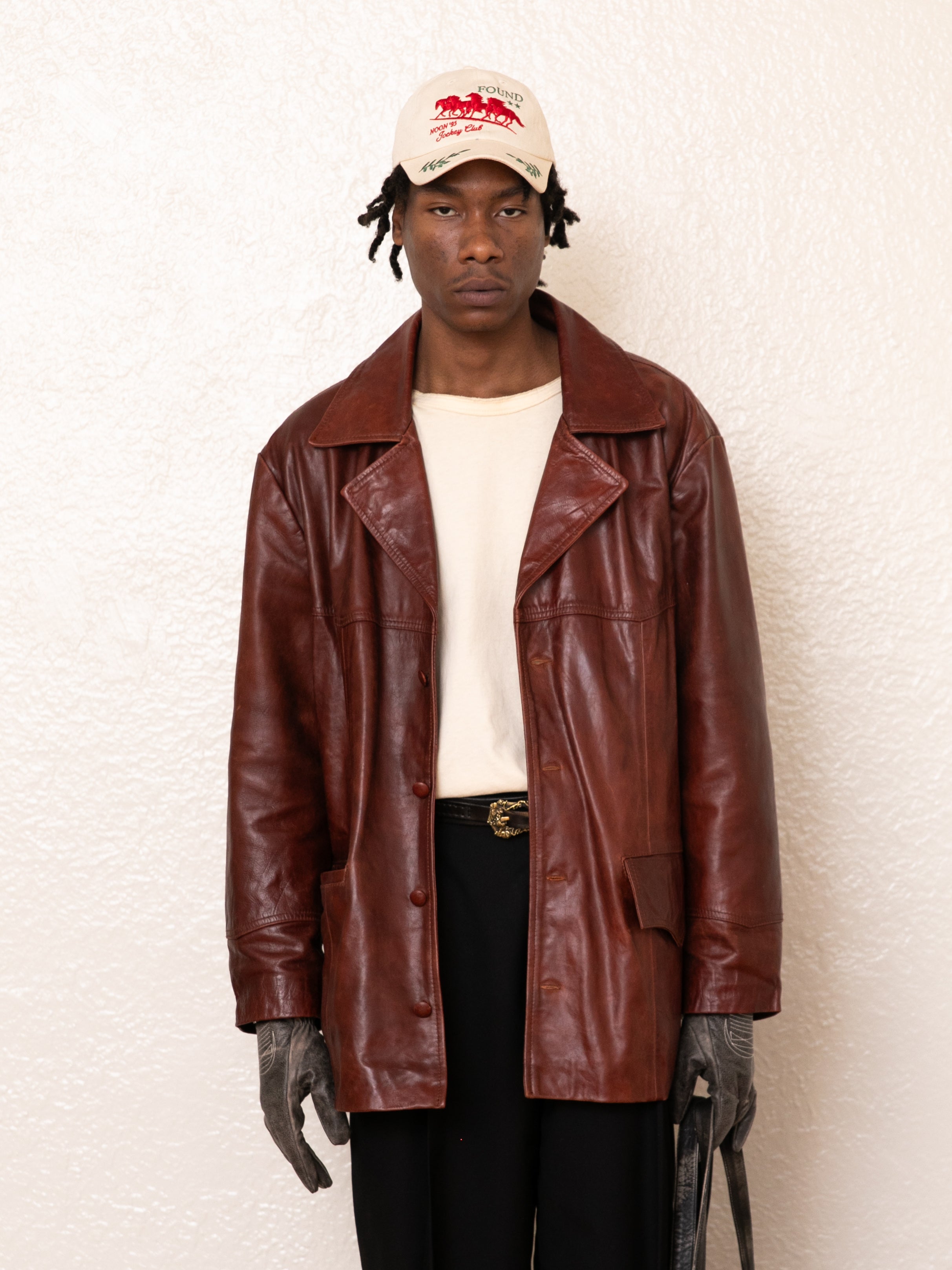 Against a white backdrop, they wear a Claret Western Leather Blazer from FOUND, paired with a beige shirt, black pants, gray gloves, and a white cap. This ensemble stylishly highlights an ethically made wardrobe choice.