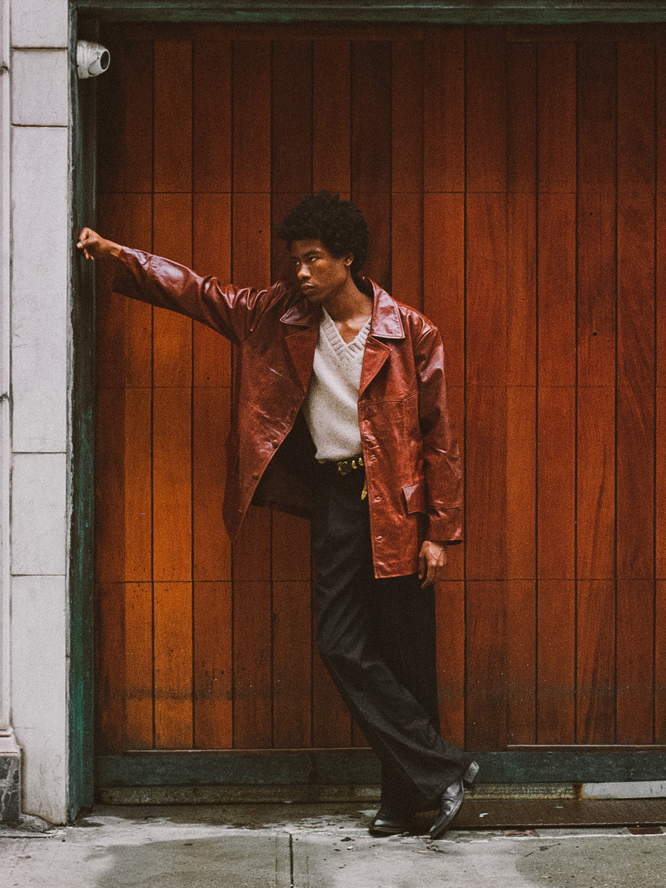 A person in a Claret Western Leather Blazer by FOUND and dark pants leans against a wooden wall, glancing to the side.