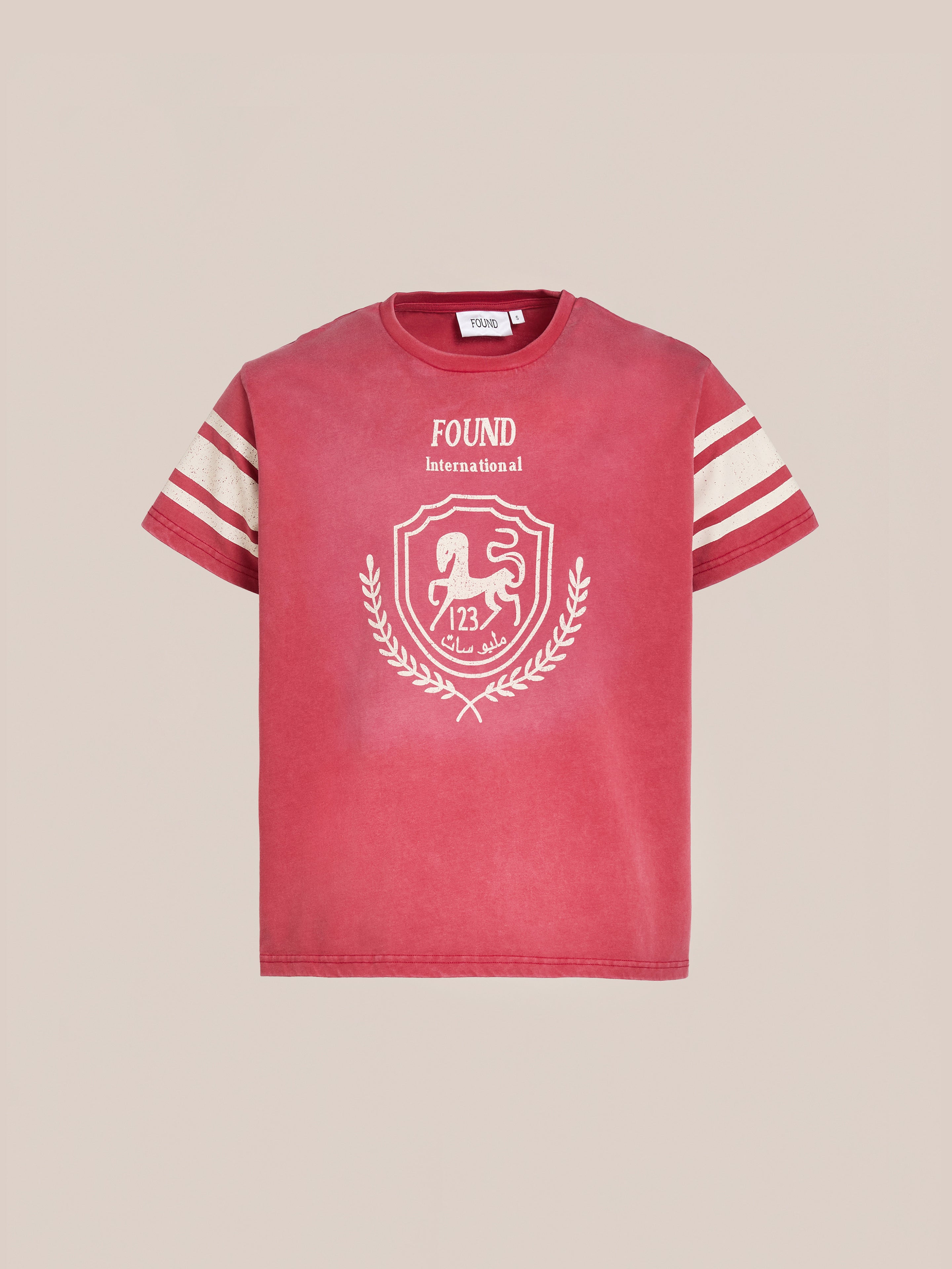 The Collegiate Crest Tee by FOUND is a red t-shirt featuring white sleeve stripes, a central lion emblem, number 123, and the text "FOUND International," blending vintage athletic charm with modern style.