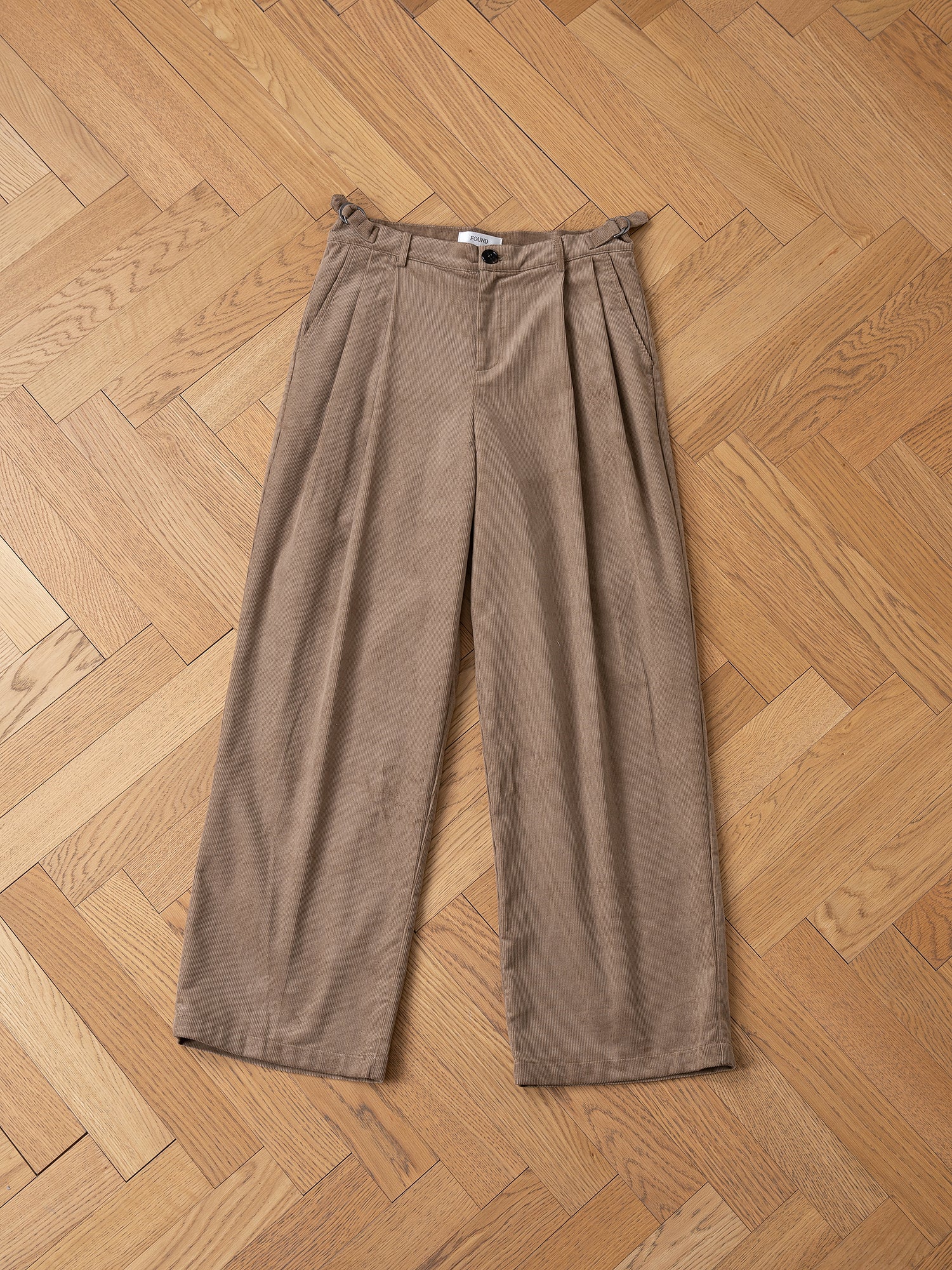 The Corduroy Pleated Trousers by FOUND, featuring a high-rise waistband with double pleats and a single button, are elegantly displayed on a wooden herringbone floor.