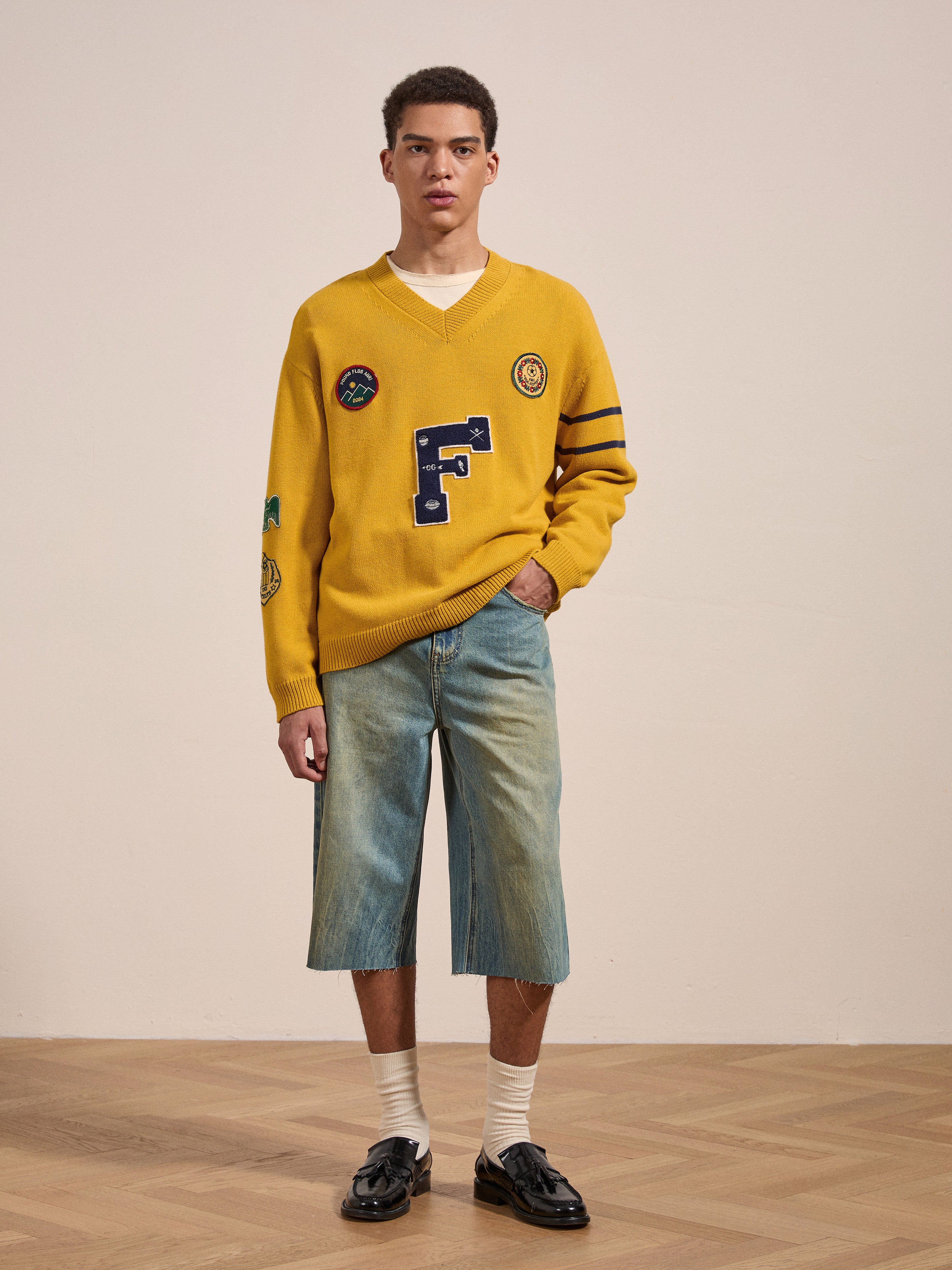 A person stands indoors wearing a yellow sweater with patches, Lacy Jean Jorts by FOUND, white socks, and black loafers. They have short curly hair and a neutral expression.