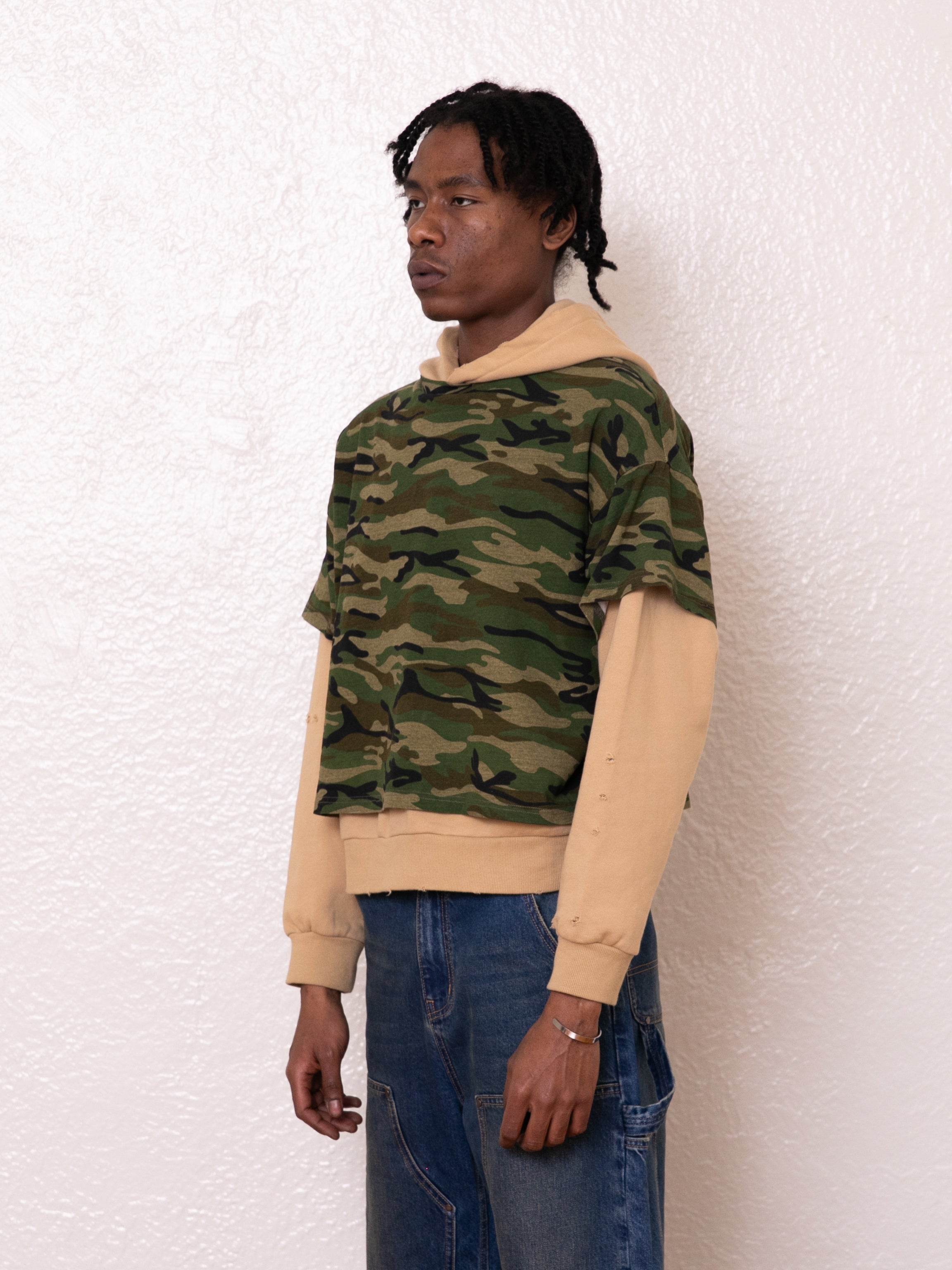 A person stands against a textured white wall, exuding a vintage look with their camouflage top layered over the FOUND Double Layer Hoodie crafted from soft French terry cotton, effortlessly paired with blue jeans.