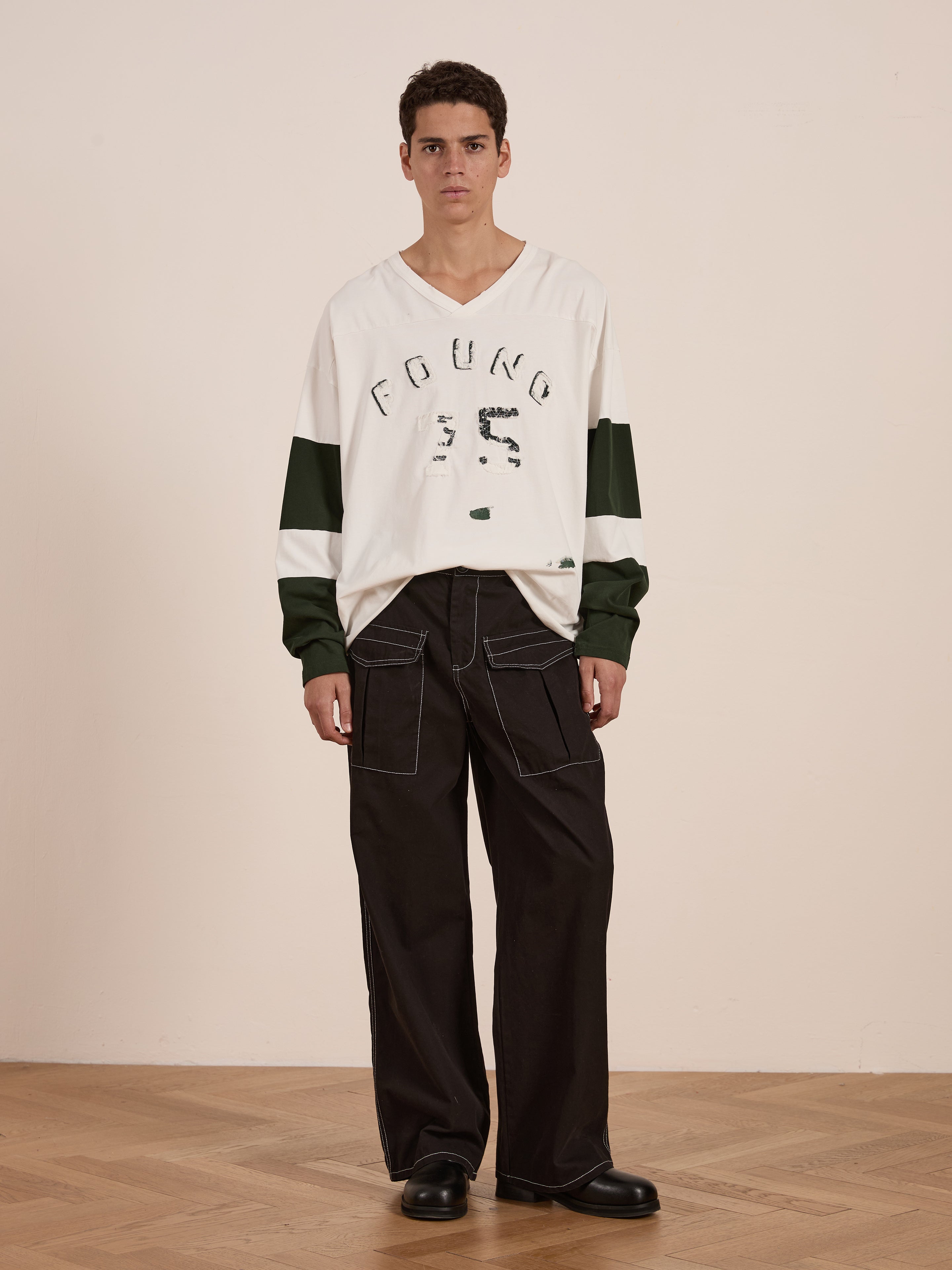 A person confidently stands on a wooden floor in a vintage sportswear ensemble, featuring the Distressed LS Team Jersey by FOUND, a white shirt with green sleeves and black wide-legged pants paired with black shoes, embodying the perfect relaxed fit look.