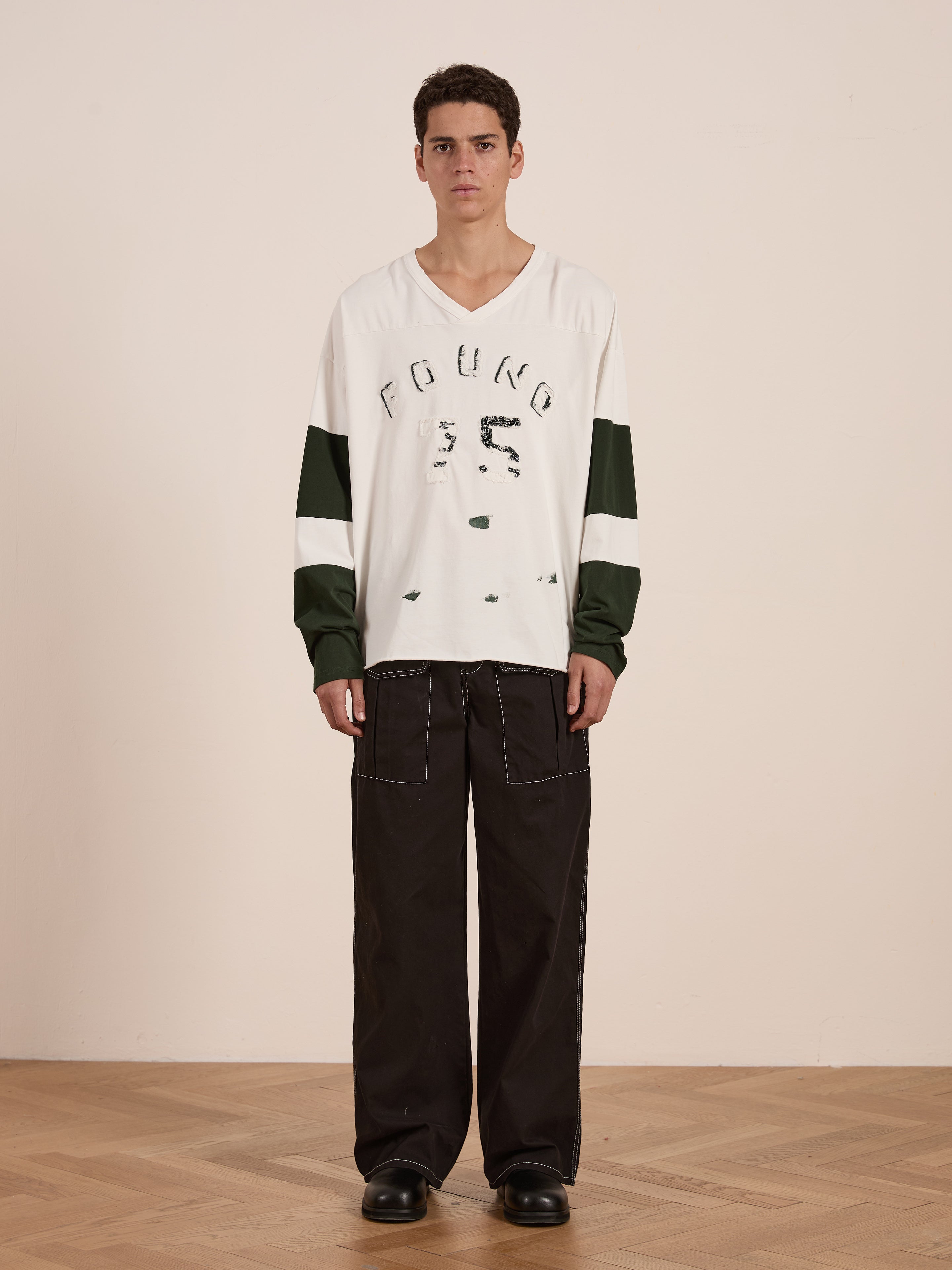 A person stands on a wooden floor wearing the Distressed LS Team Jersey by FOUND, a relaxed-fit shirt in white and green with the brand name, paired with black pants featuring white stitching, creating a vintage sportswear vibe.