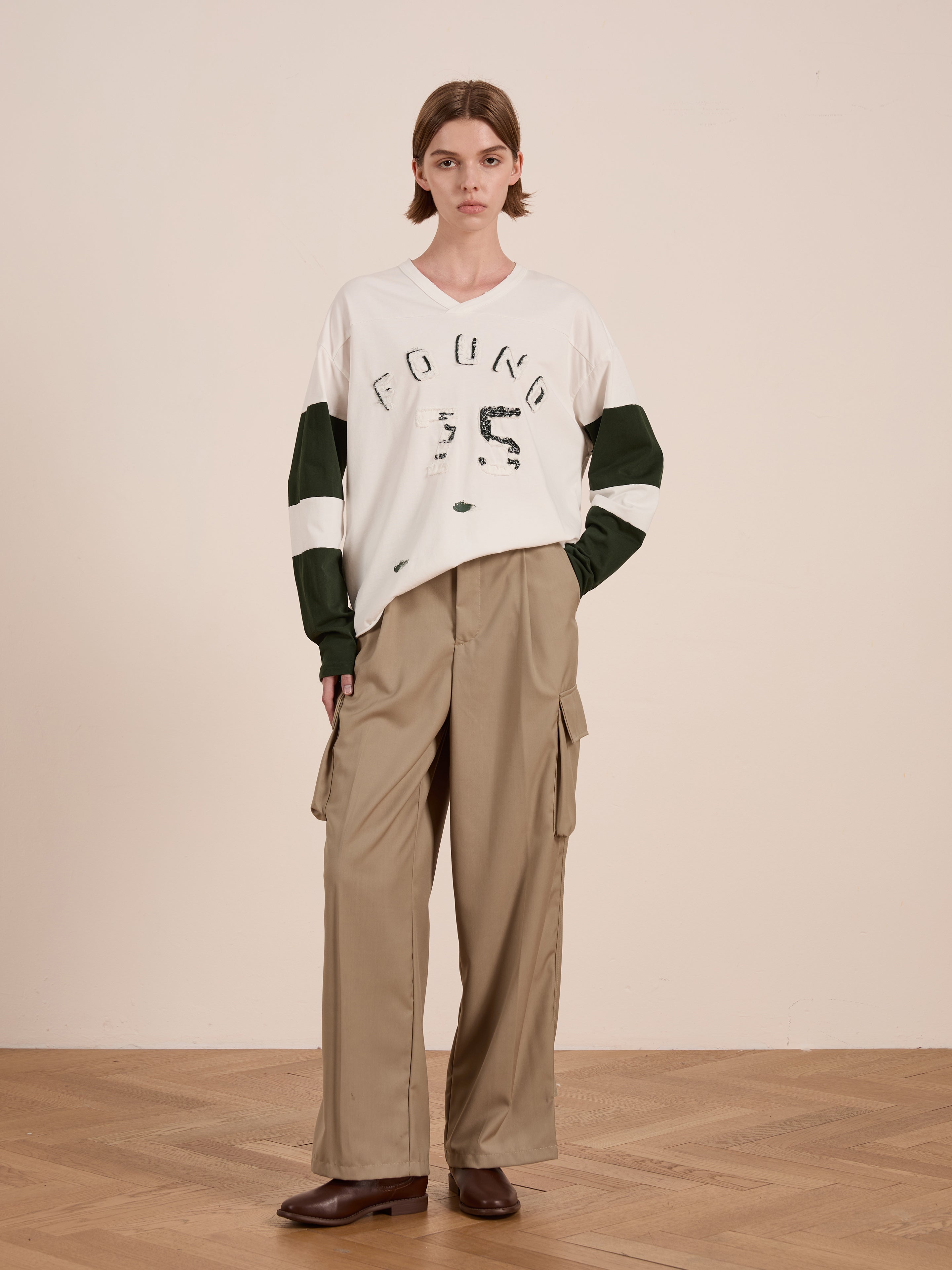 Indoors, a person wears a Distressed LS Team Jersey by FOUND, paired with beige wide-leg cargo pants and brown boots, creating a laid-back look against a plain wall.