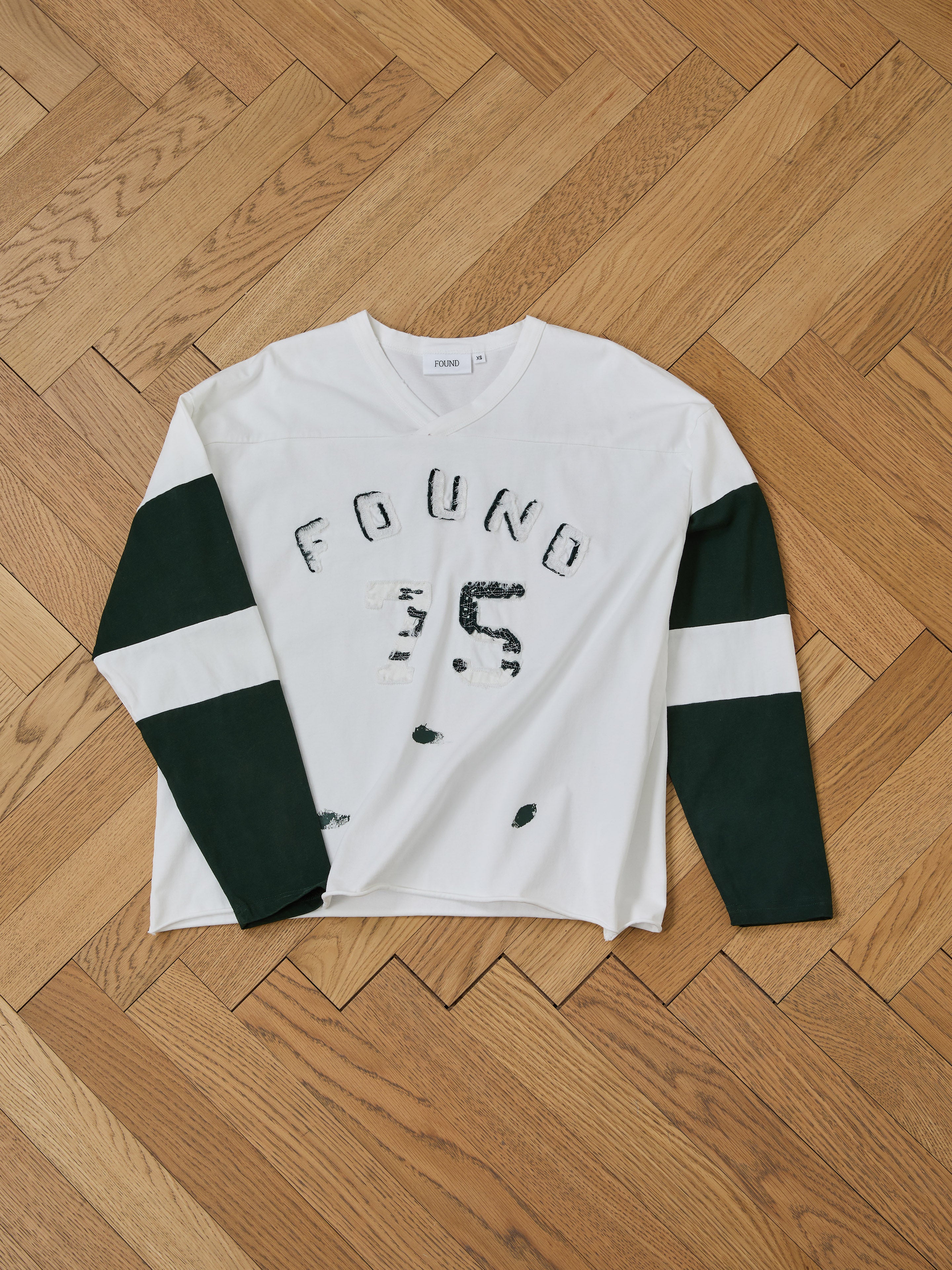 The Distressed LS Team Jersey by FOUND is a white long-sleeve shirt with green sleeves and "FOUND 95" on the front, featuring a relaxed fit inspired by vintage sportswear, displayed on a wooden floor.