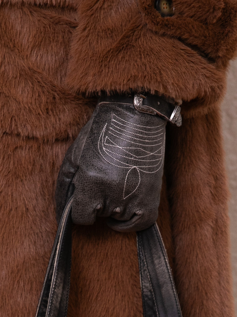 A close-up reveals a gloved hand adorned with stitched patterns, gripping a leather strap while wearing a brown furry coat. The Distressed Leather Western Gloves by FOUND contribute an authentic Western flair to the ensemble.